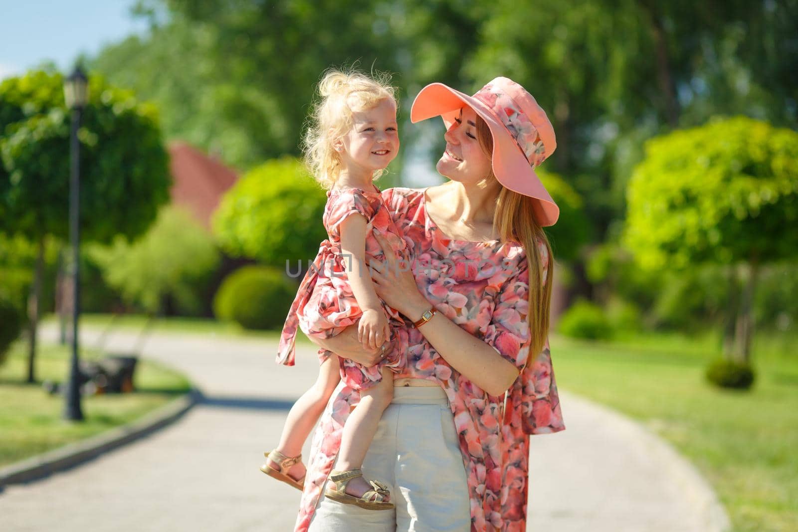 A charming girl in a light summer sundress walks in a green park with her little daughter, holding her in her arms. Enjoys warm sunny summer days.