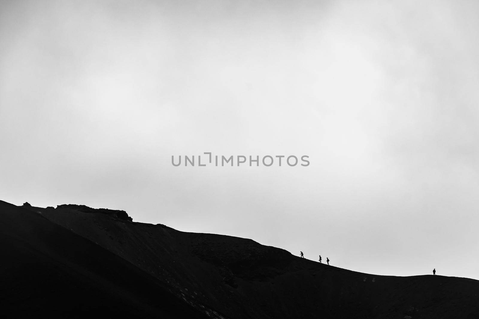 Minimalist image of small silhouette of men climbing on the edge of Etna volcano crater with a cloudy background