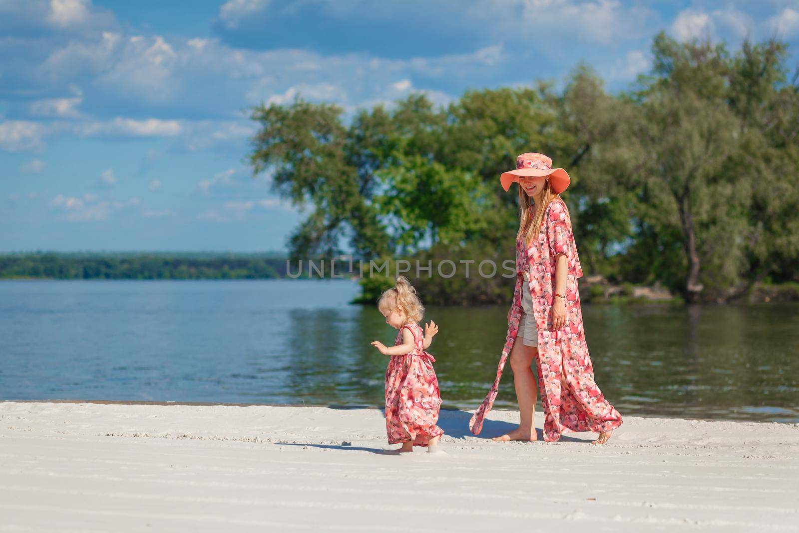 A charming girl in a light summer sundress walks on the sandy beach with her little daughter. Enjoys warm sunny summer days by Try_my_best