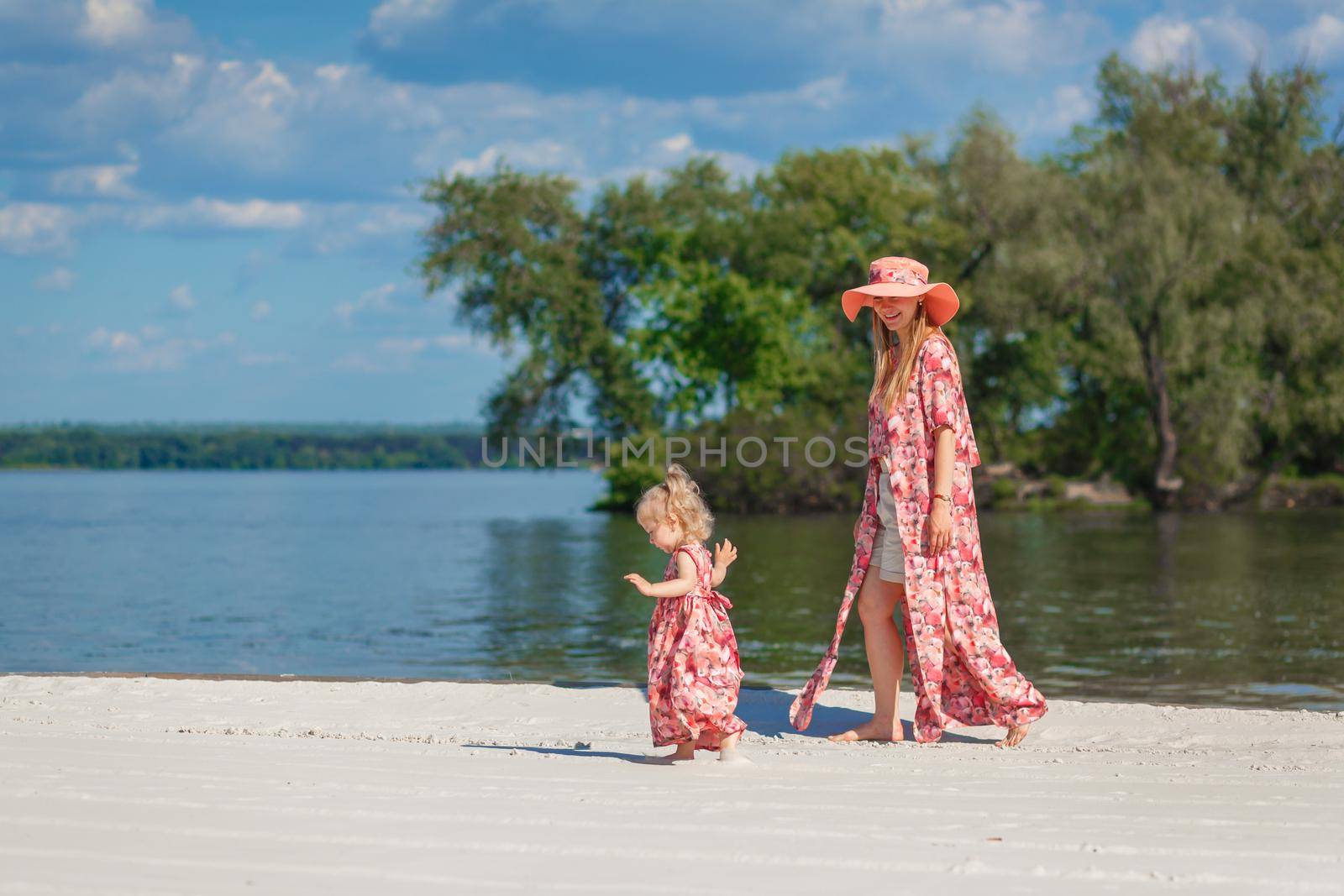 A charming girl in a light summer sundress walks on the sandy beach with her little daughter. Enjoys warm sunny summer days.