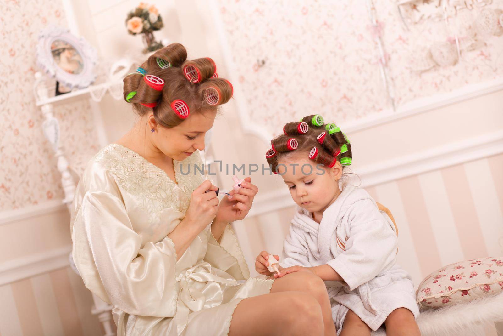 Adorable little girl with her mother in curlers paint their fingernails. Copies mom's behavior. Mom teaches her daughter to take care of herself. Beauty day.