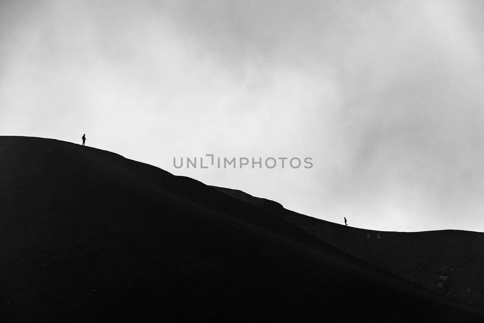 Minimalist image of small silhouette of men climbing on the edge of Etna volcano crater with a cloudy background