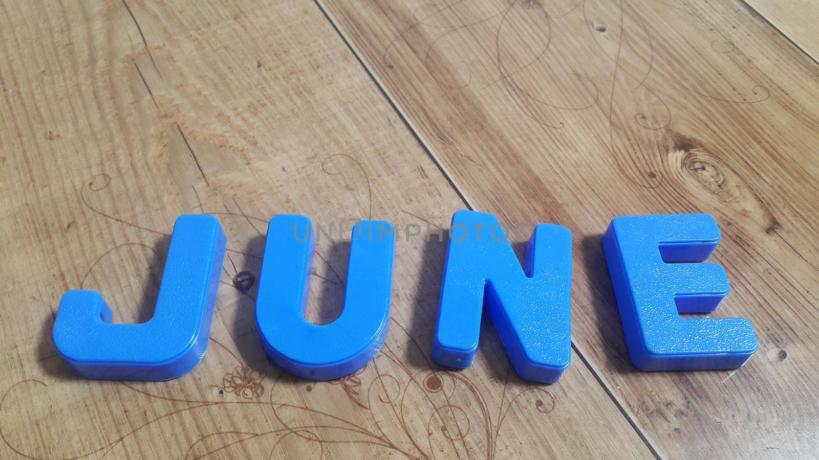 Plastic colored alphabets making word June are placed on a wooden floor. These plastic letters can be used for teaching kids.