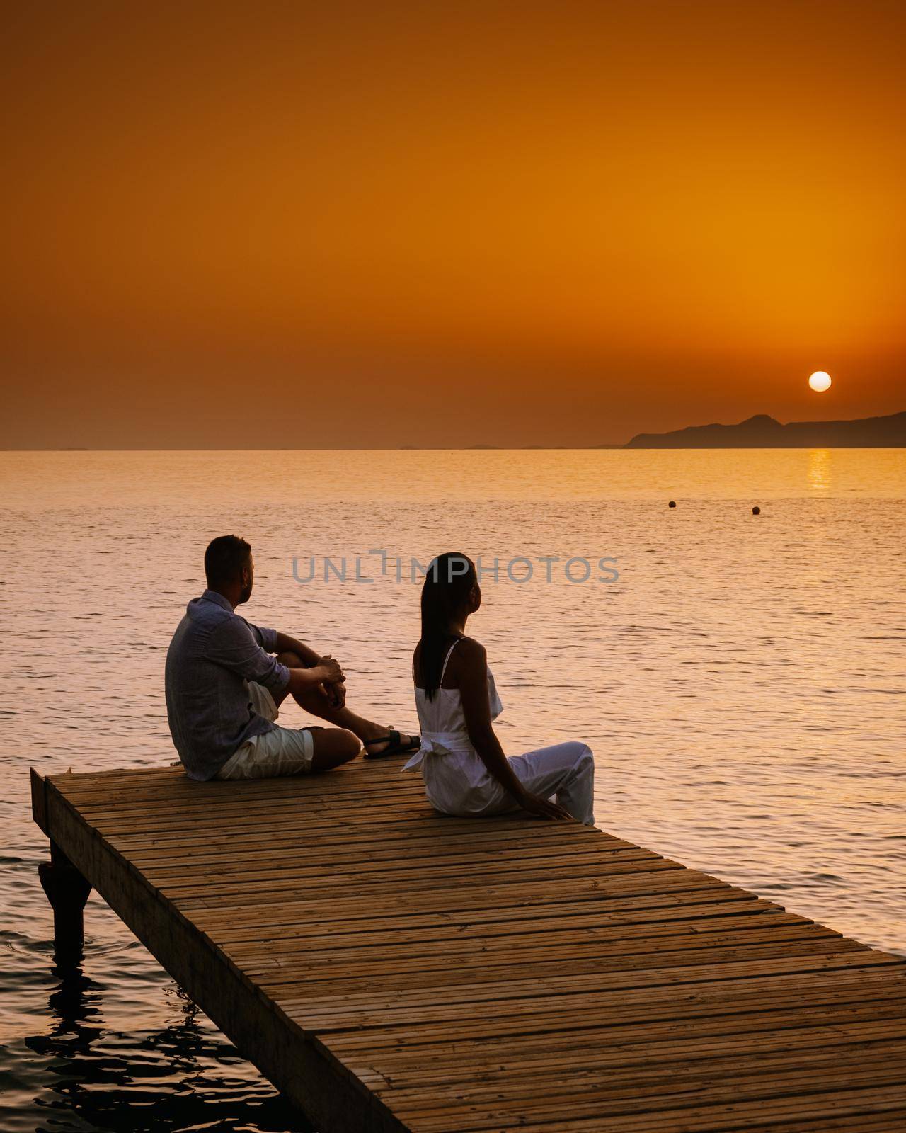 young romantic couple in love is sitting and hugging on wooden pier at the beach in sunrise time with golden sky. Vacation and travel concept. Romantic young couple dating at seaside. Crete Greece