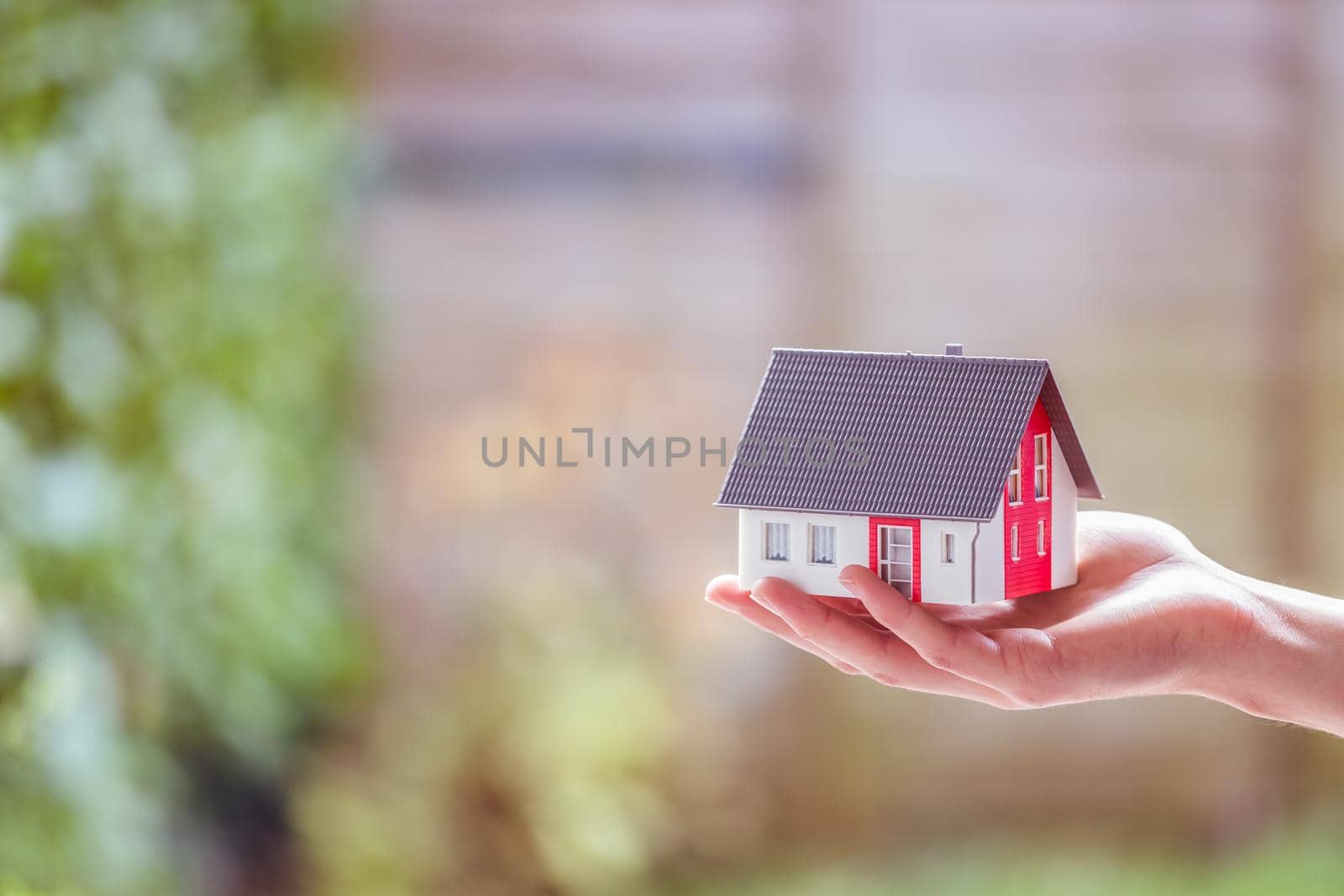 Adult hand is holding red house model, outdoors. Concept for new home, property and estate by Daxenbichler