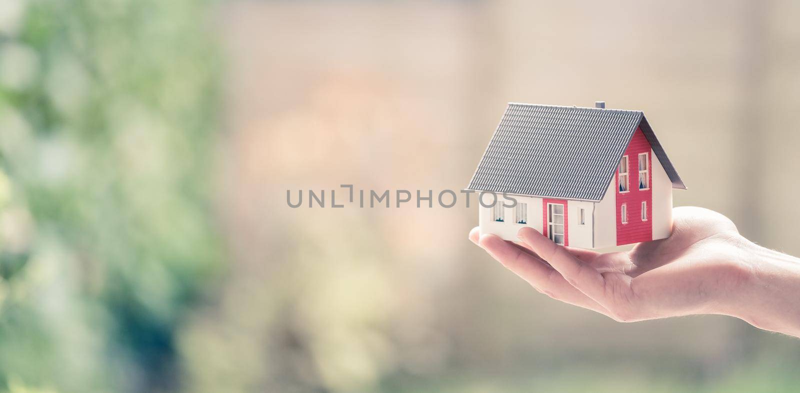 Adult hand is holding red house model, outdoors. Concept for new home, property and estate. Text space. by Daxenbichler
