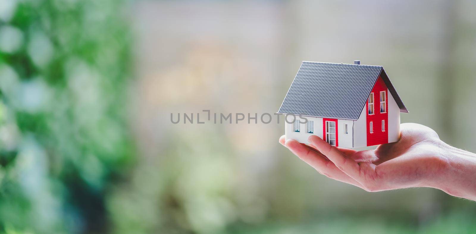 Adult hand is holding red house model, outdoors. Concept for new home, property and estate. Text space. by Daxenbichler