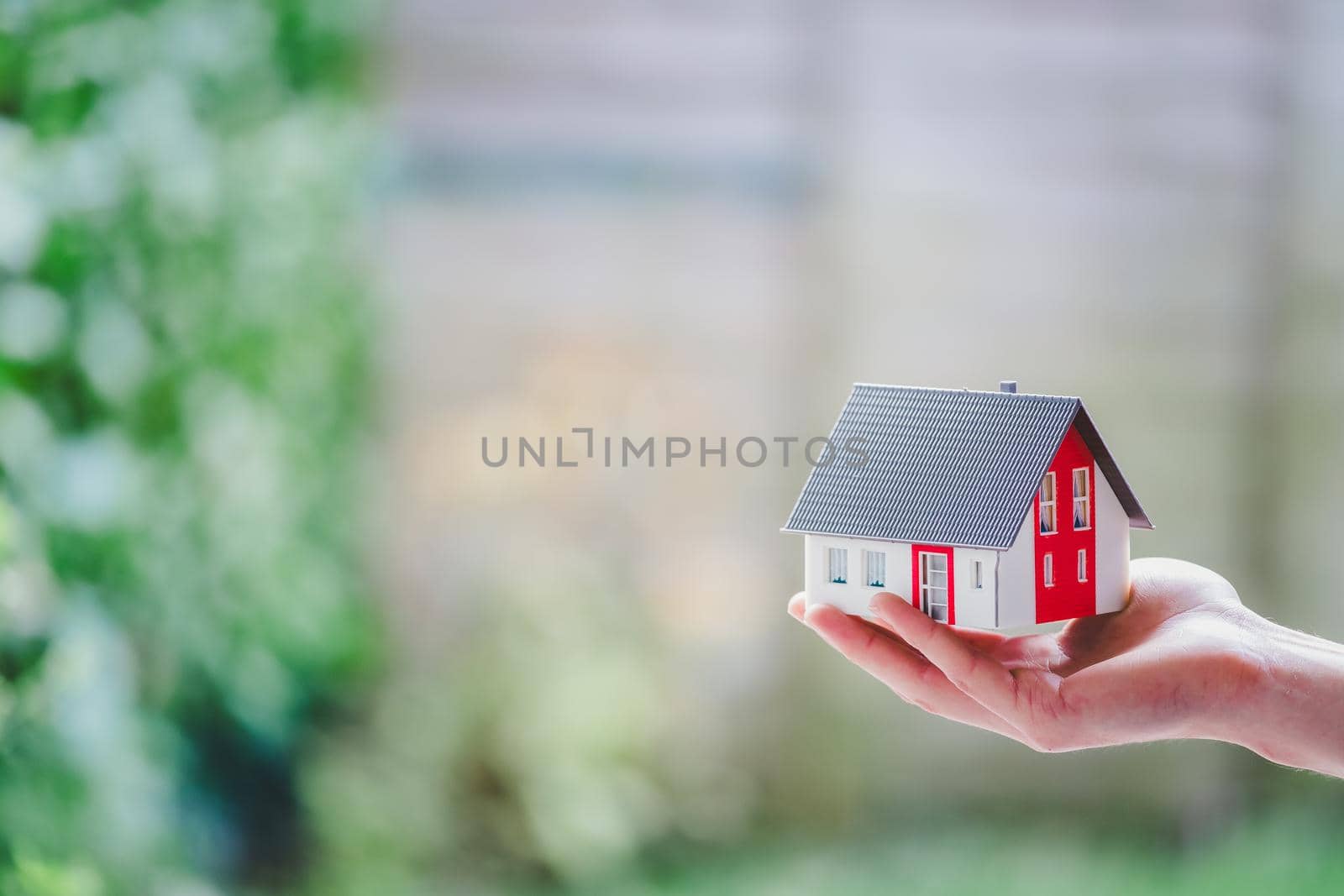 Adult hand is holding red house model, outdoors. Concept for new home, property and estate by Daxenbichler