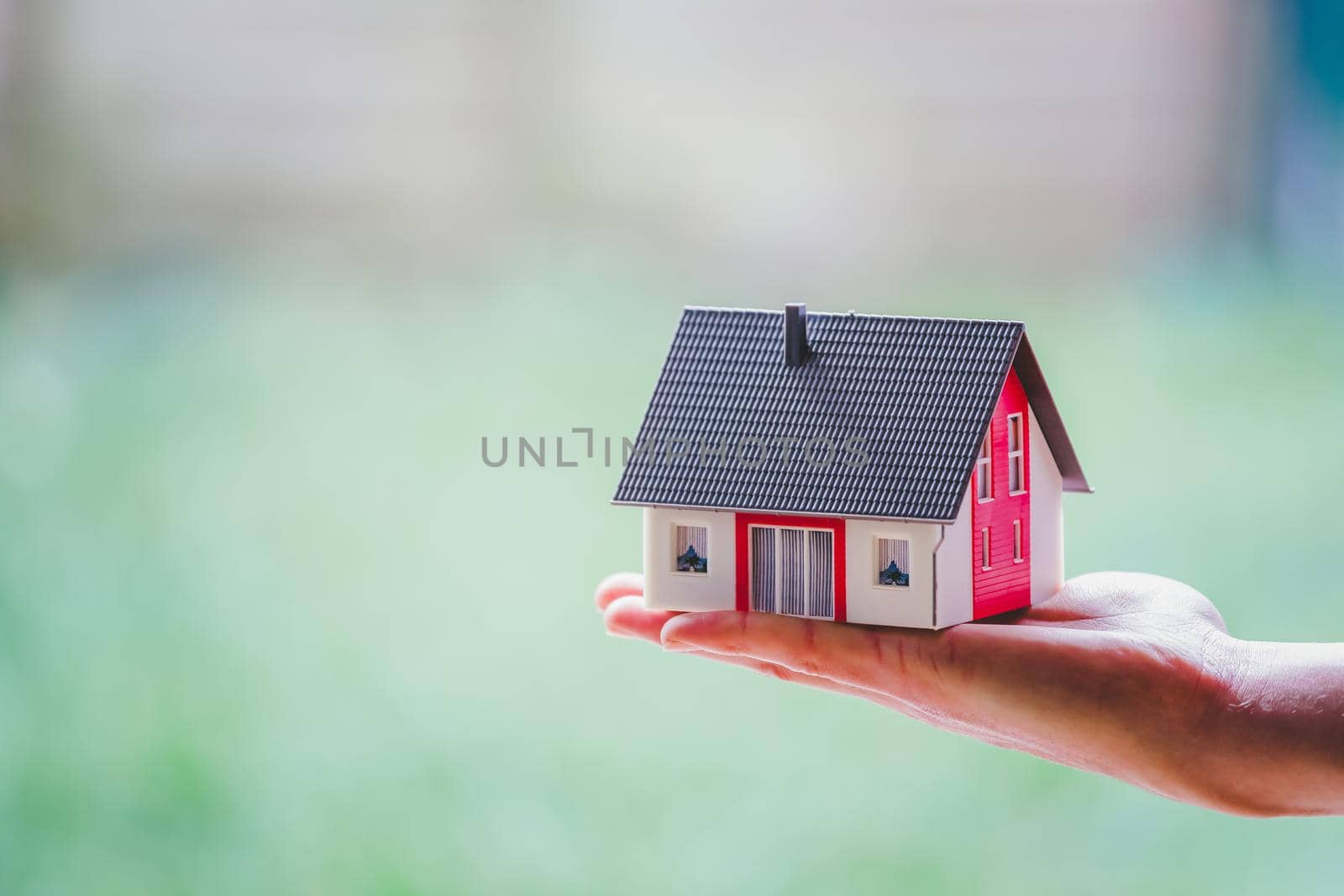 Adult hand is holding red house model, outdoors. Concept for new home, property and estate by Daxenbichler