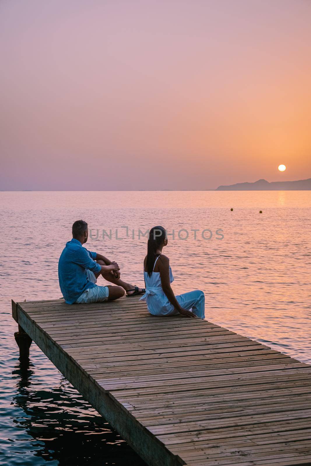 Crete Greece, young romantic couple in love is sitting and hugging on wooden pier at the beach in sunrise time with golden sky. Vacation and travel concept. Romantic young couple dating at seaside by fokkebok