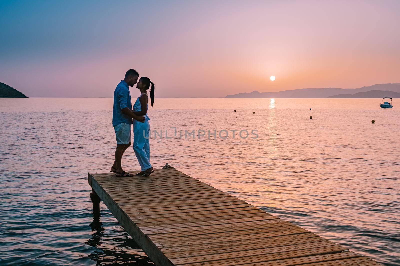 young romantic couple in love is sitting and hugging on wooden pier at the beach in sunrise time with golden sky. Vacation and travel concept. Romantic young couple dating at seaside. Crete Greece