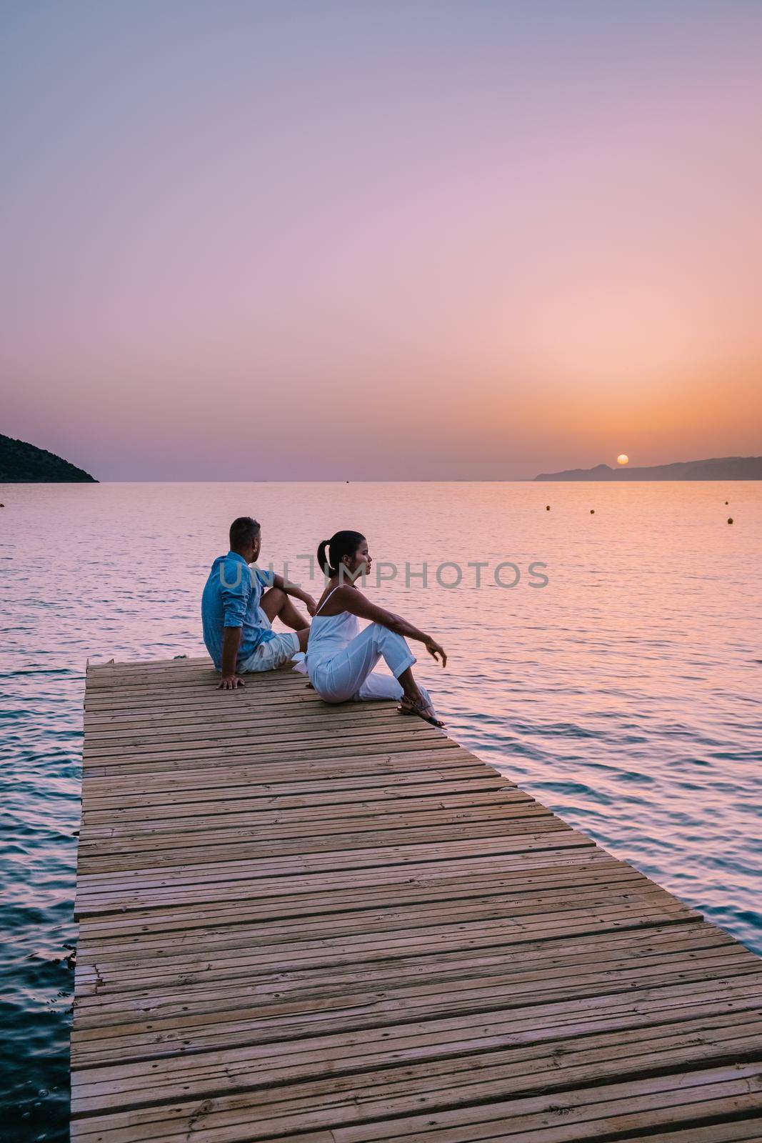 Crete Greece, young romantic couple in love is sitting and hugging on wooden pier at the beach in sunrise time with golden sky. Vacation and travel concept. Romantic young couple dating at seaside by fokkebok