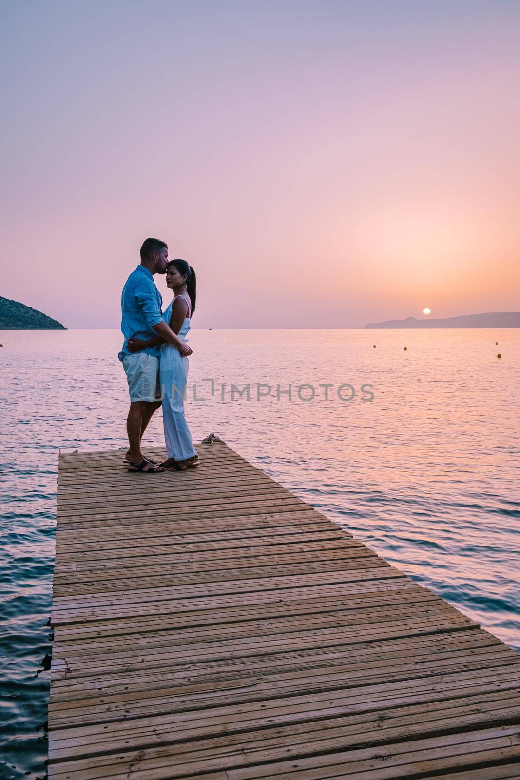 Crete Greece, young romantic couple in love is sitting and hugging on wooden pier at the beach in sunrise time with golden sky. Vacation and travel concept. Romantic young couple dating at seaside by fokkebok