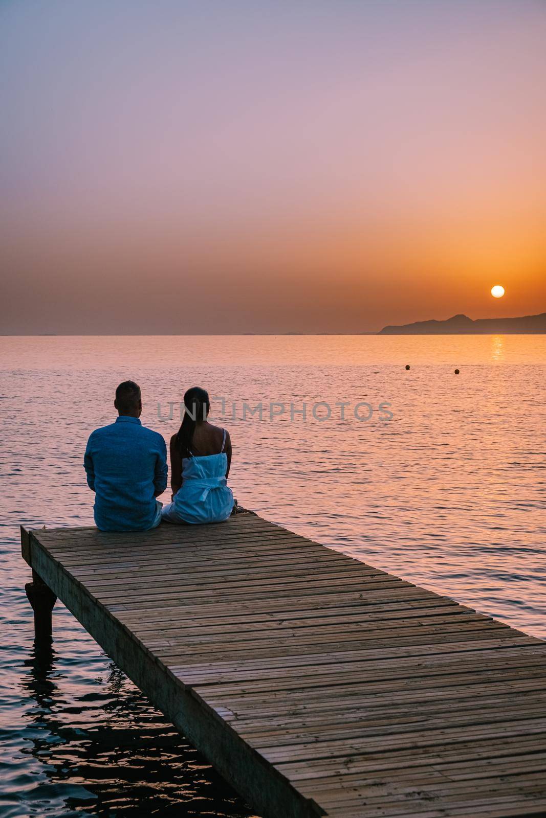 Crete Greece, young romantic couple in love is sitting and hugging on wooden pier at the beach in sunrise time with golden sky. Vacation and travel concept. Romantic young couple dating at seaside by fokkebok