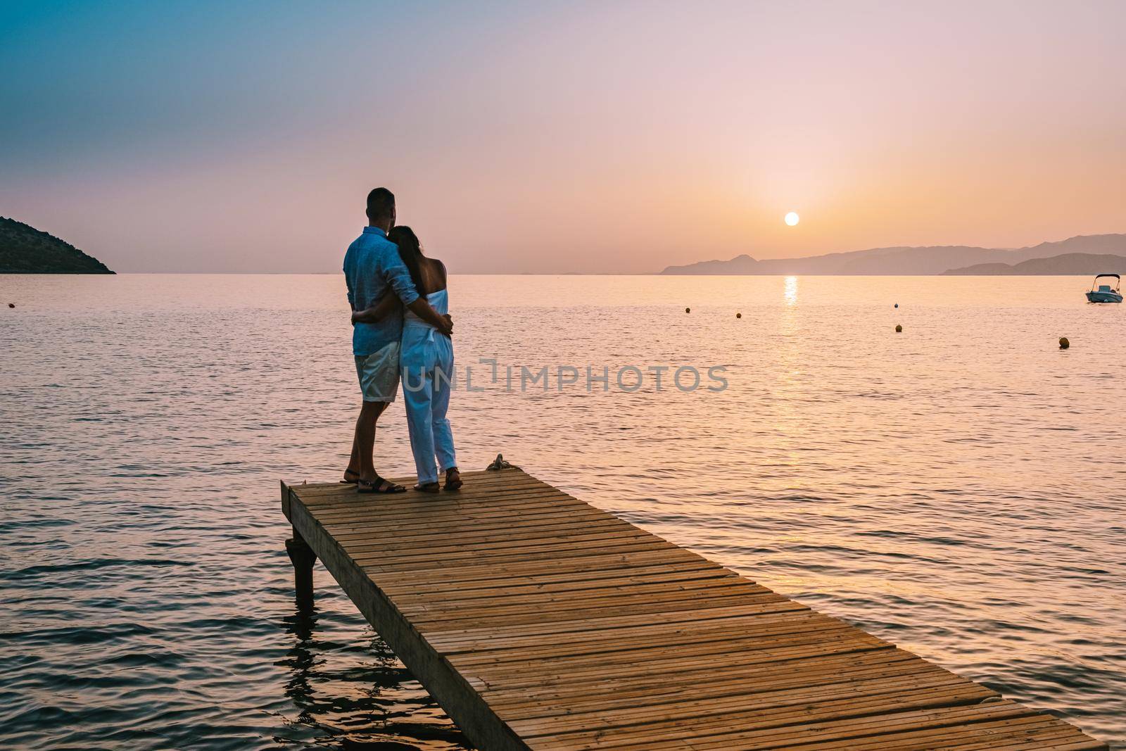 young romantic couple in love is sitting and hugging on wooden pier at the beach in sunrise time with golden sky. Vacation and travel concept. Romantic young couple dating at seaside. Crete Greece