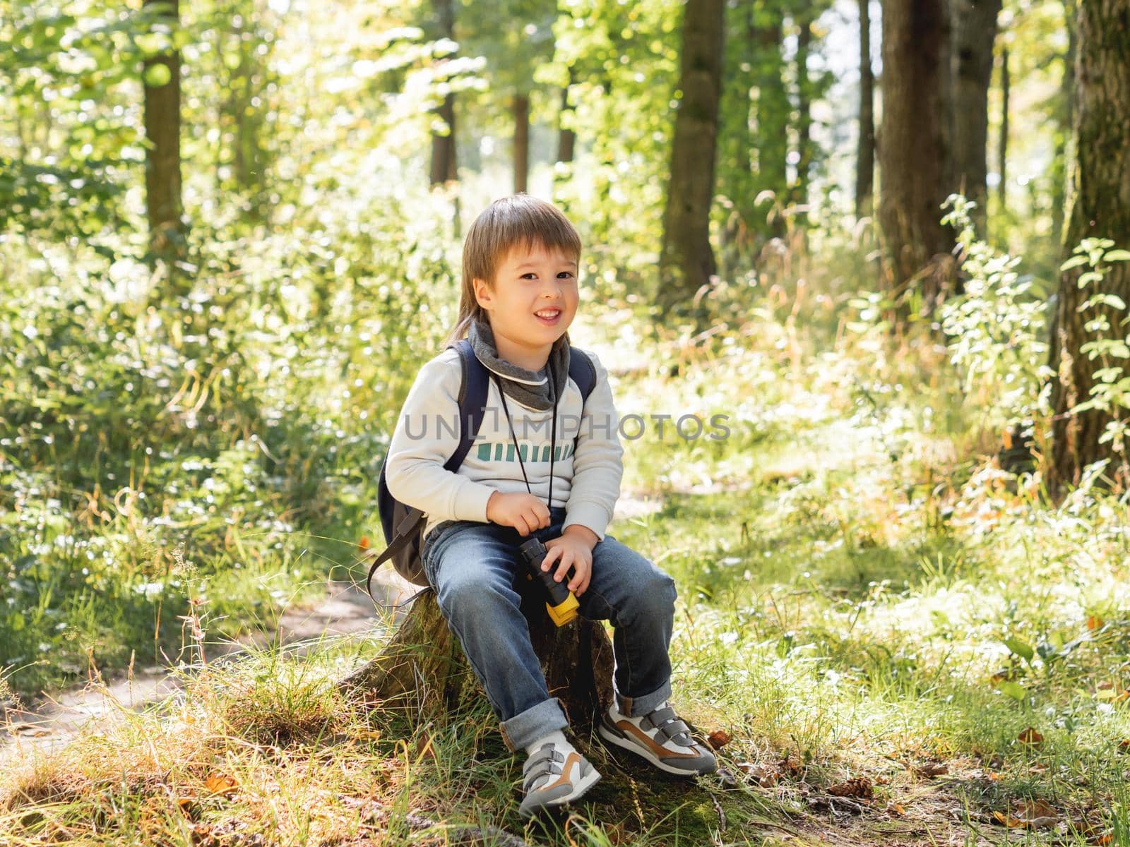 Little explorer on hike in forest. Boy with binoculars sits on stump. Outdoor leisure activity for children. Summer journey for young tourist. by aksenovko