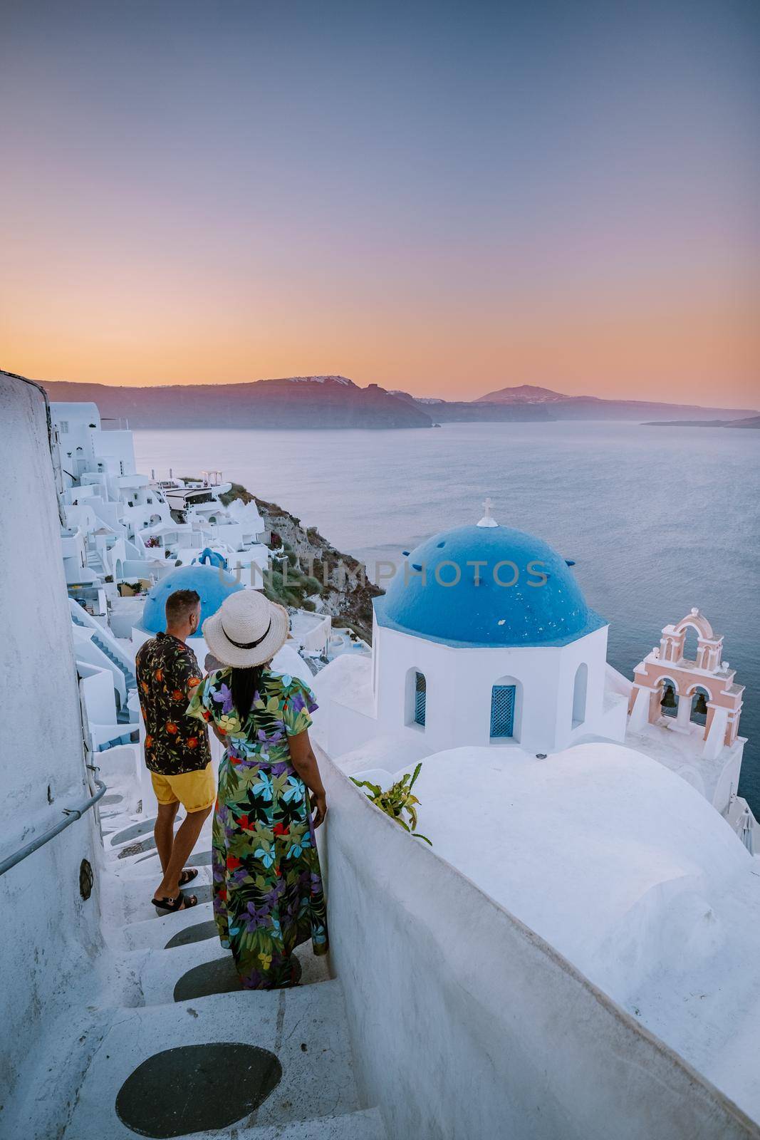 Santorini Greece, young couple on luxury vacation at the Island of Santorini watching sunrise by the blue dome church and whitewashed village of Oia Santorini Greece during sunrise during summer vacation, men and woman on holiday in Greece