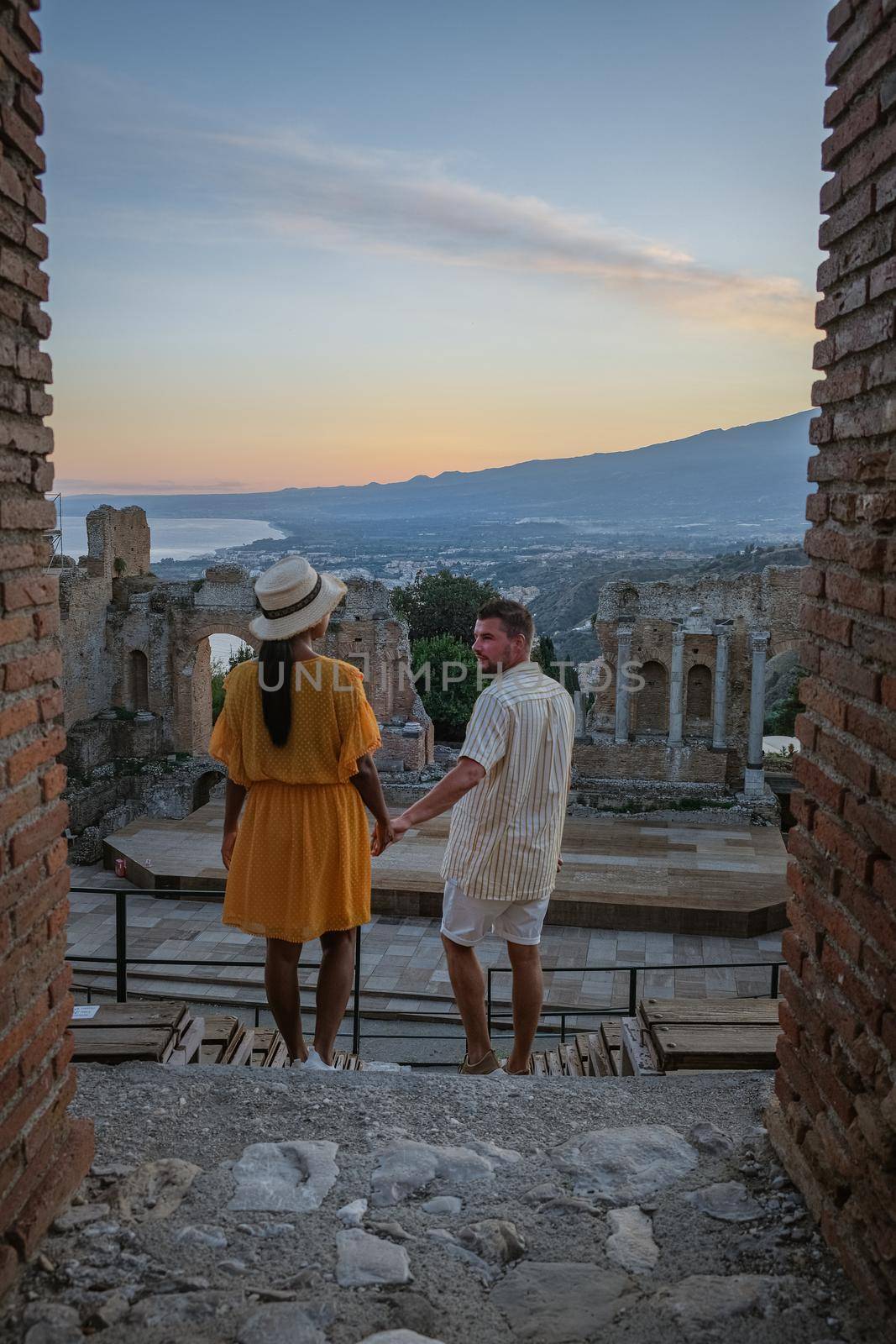 couple men and woman visit Ruins of Ancient Greek theatre in Taormina on background of Etna Volcano, Italy. Taormina located in Metropolitan City of Messina, on east coast of island of Sicily. by fokkebok