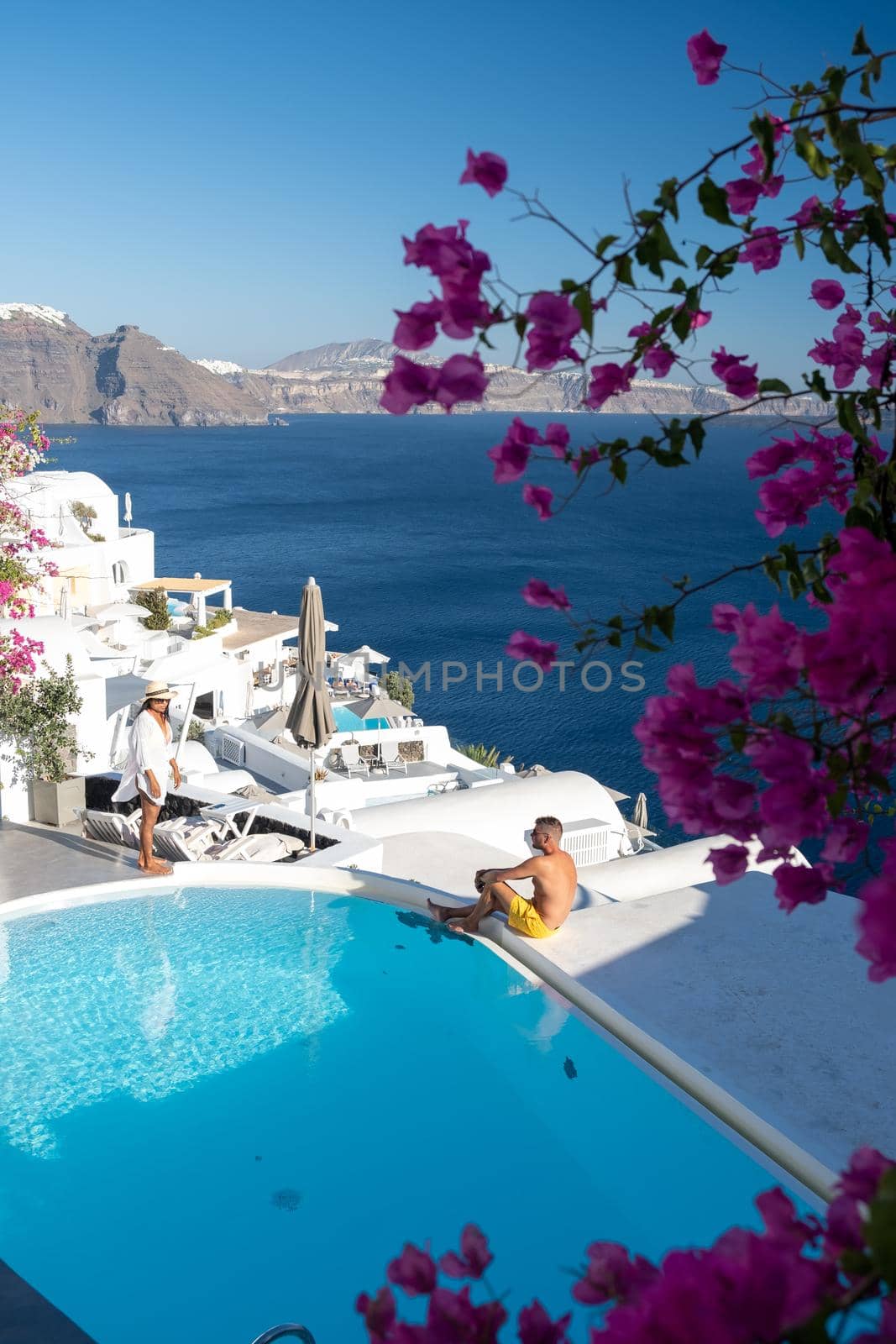 Couple at luxury infinity pool, antorini Greece, young couple on luxury vacation at the Island of Santorini watching sunrise by the blue dome church and whitewashed village of Oia Santorini Greece  by fokkebok