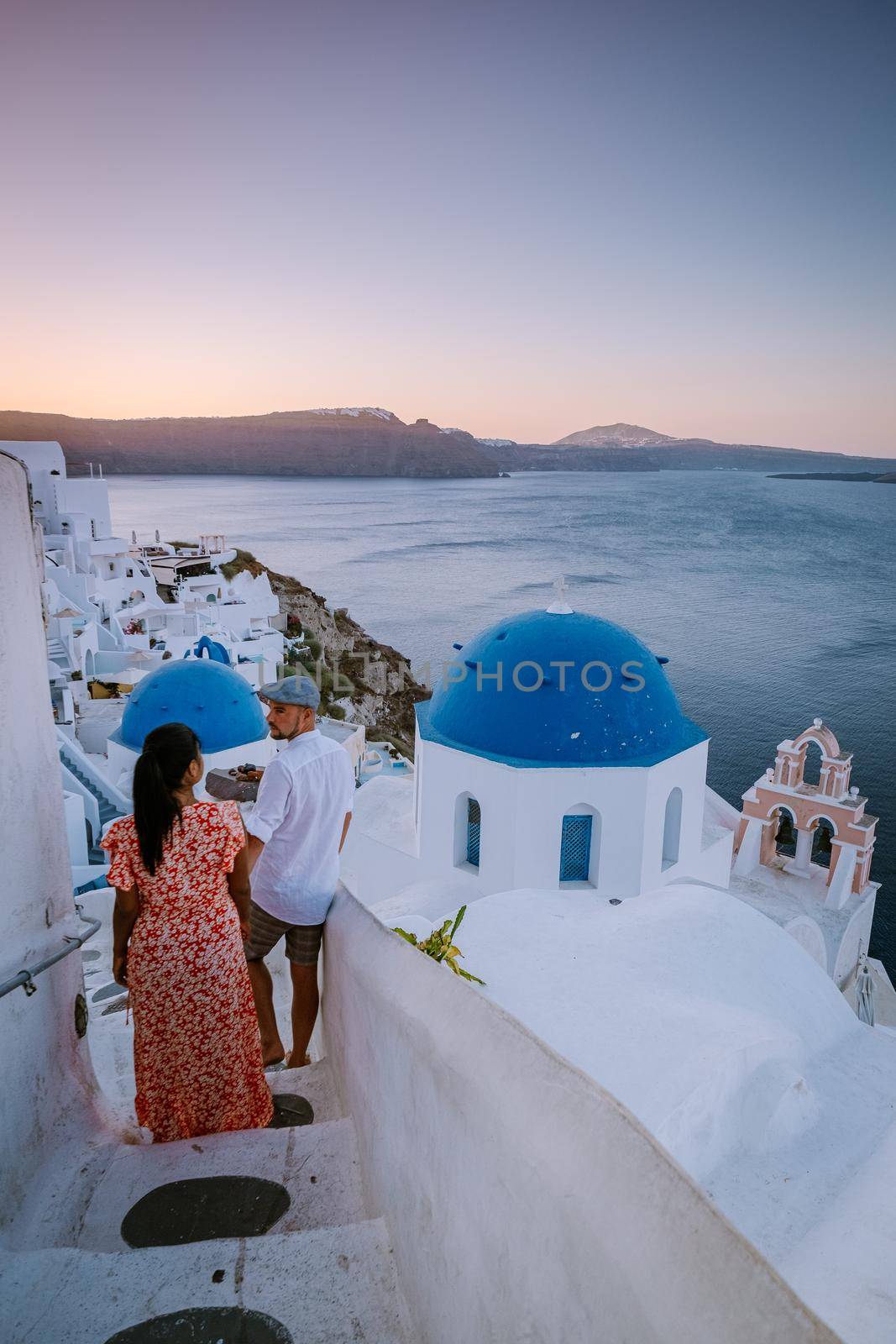 Santorini Greece, young couple on luxury vacation at the Island of Santorini watching sunrise by the blue dome church and whitewashed village of Oia Santorini Greece during sunrise during summer vacation, men and woman on holiday in Greece