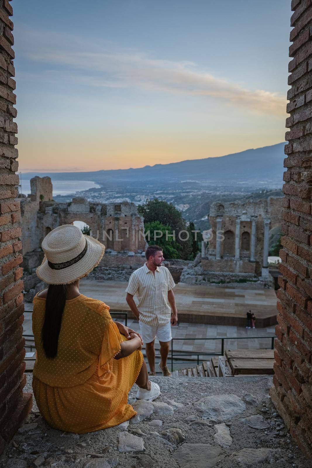 couple men and woman visit Ruins of Ancient Greek theatre in Taormina on background of Etna Volcano, Italy. Taormina located in Metropolitan City of Messina, on east coast of island of Sicily. by fokkebok