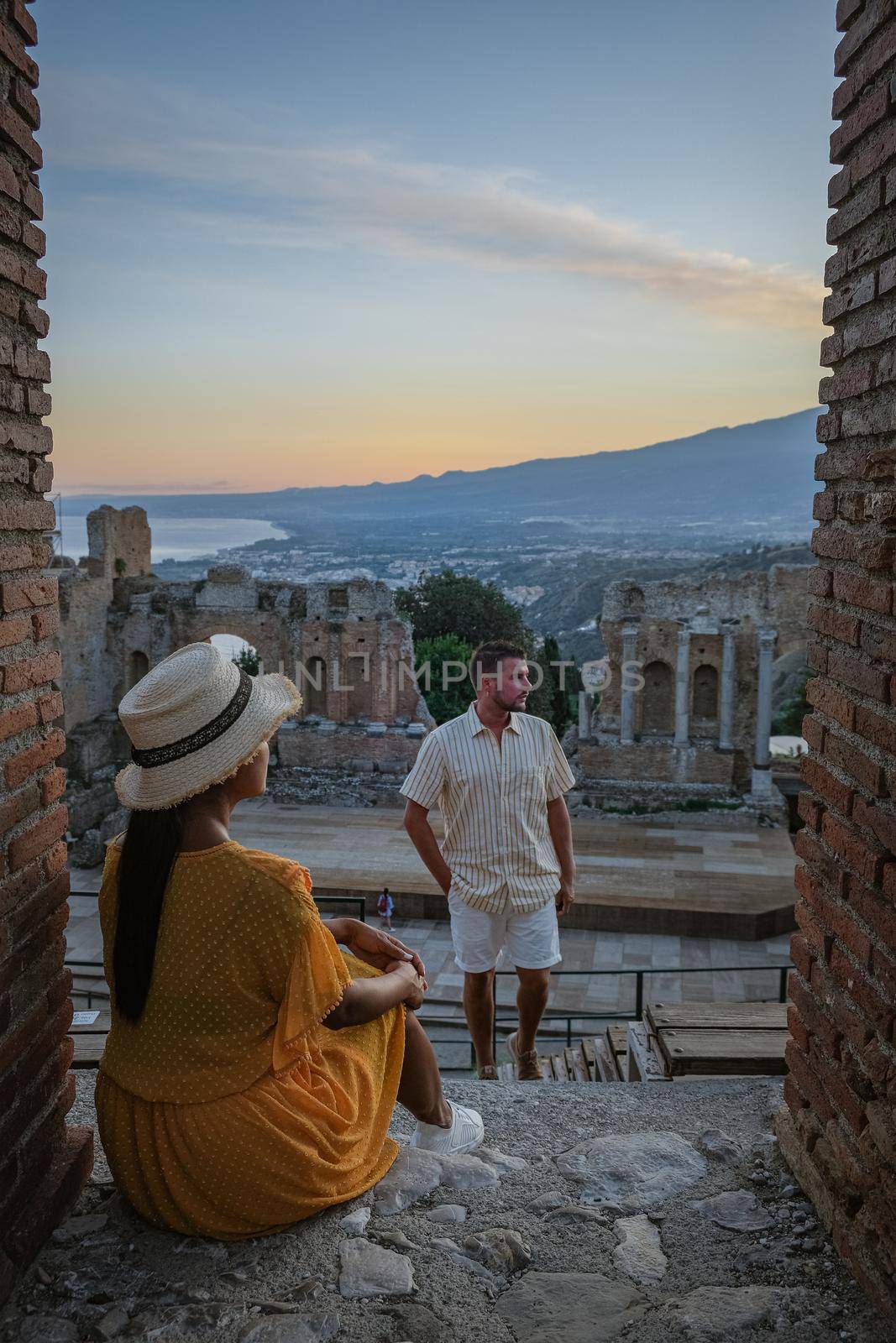 couple men and woman visit Ruins of Ancient Greek theatre in Taormina on background of Etna Volcano, Italy. Taormina located in Metropolitan City of Messina, on east coast of island of Sicily. by fokkebok