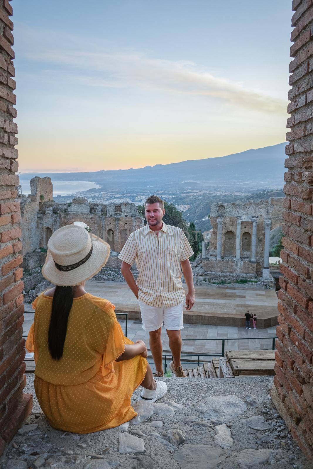 couple men and woman visit Ruins of Ancient Greek theatre in Taormina on background of Etna Volcano, Italy. Taormina located in Metropolitan City of Messina, on east coast of island of Sicily. by fokkebok