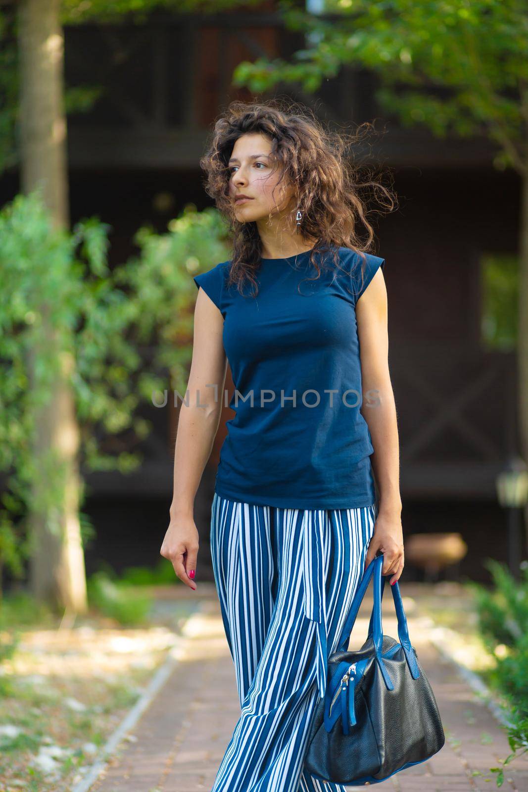 young beautiful brunette with curly hair, in a stylish outfit with a bag. Enjoying a bright summer day in a green park.