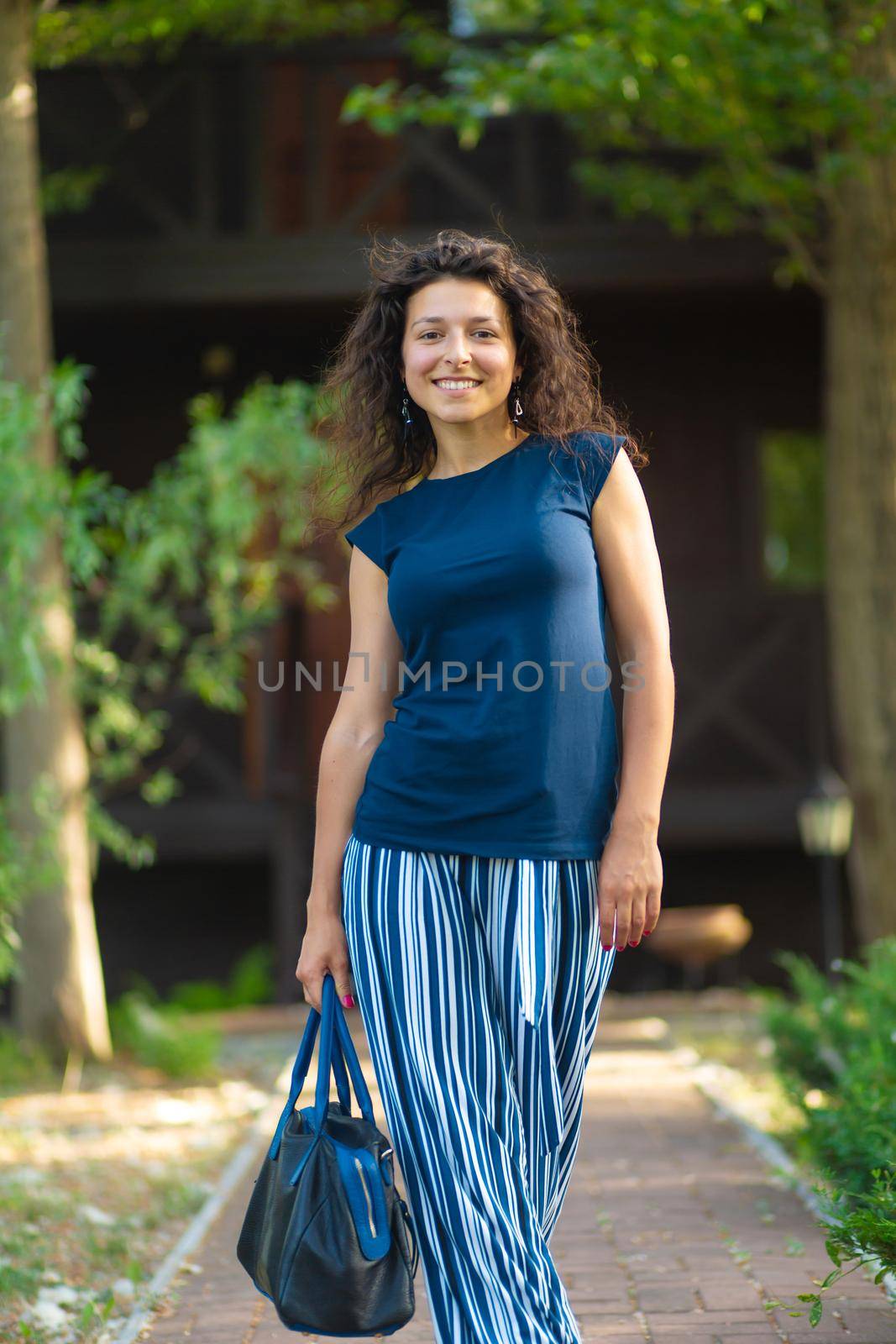 young beautiful brunette with curly hair, in a stylish outfit with a bag. Enjoying a bright summer day in a green park.