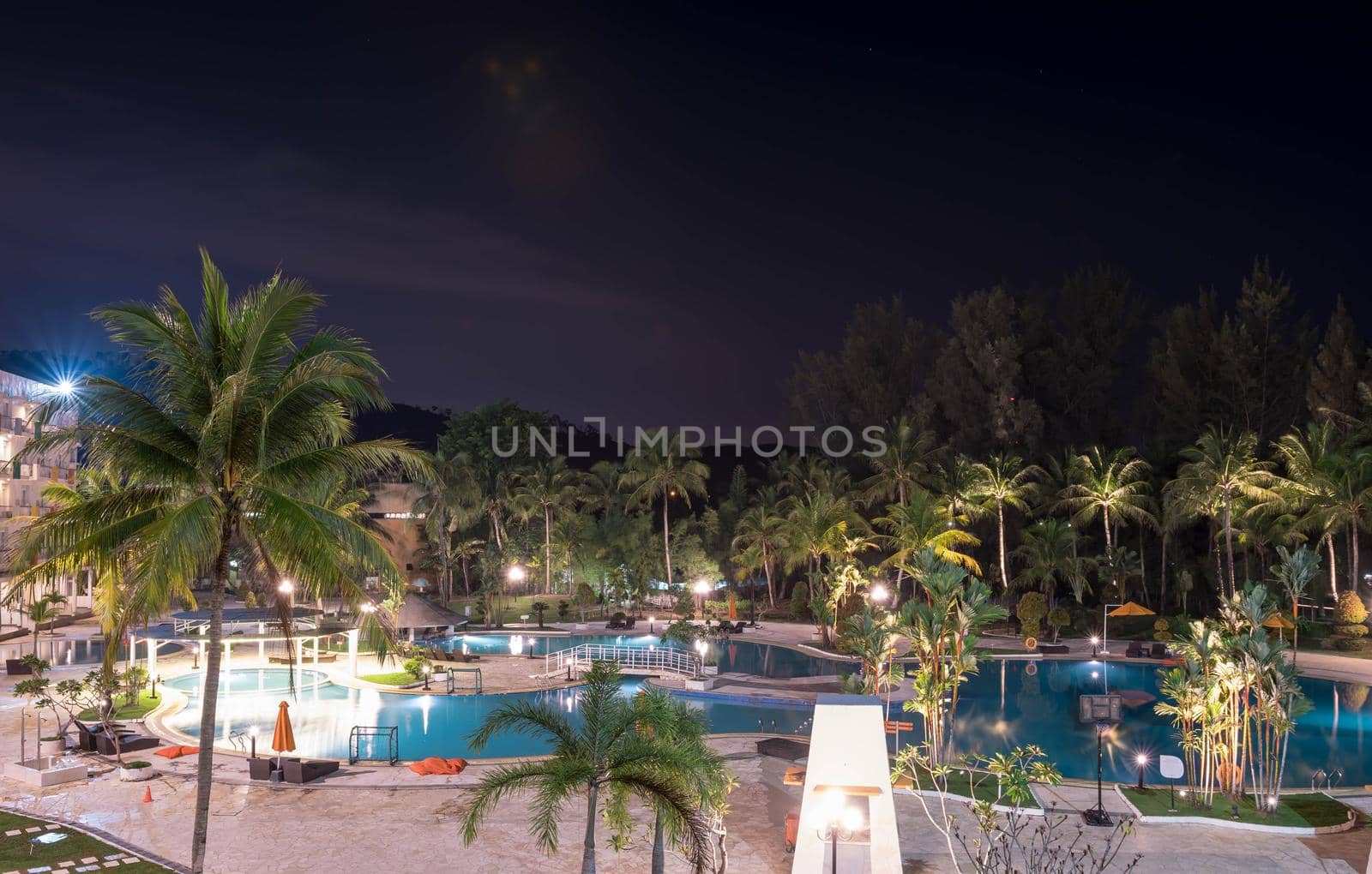 Swimming pool area in a beach hotel resort during night with trees and smooth blue swimming pool water reflecting lights long exposure somewhere in Asia