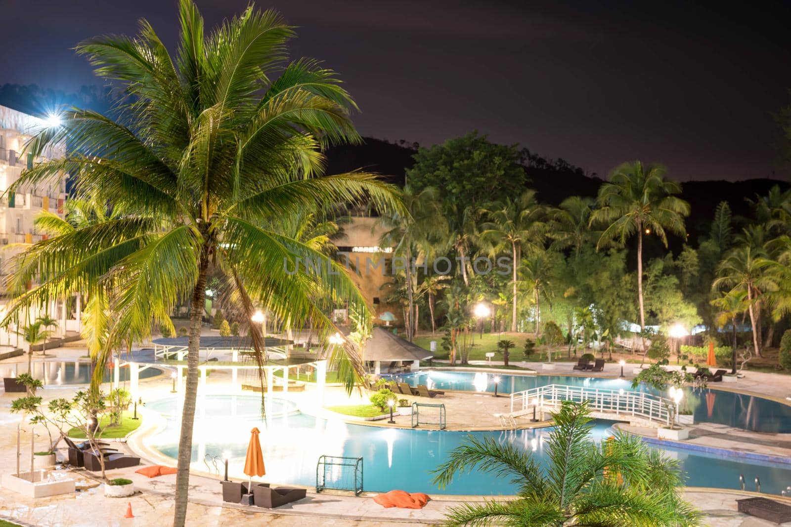 Swimming pool area in a beach hotel resort during night with trees and smooth blue swimming pool water reflecting lights long exposure somewhere in Asia