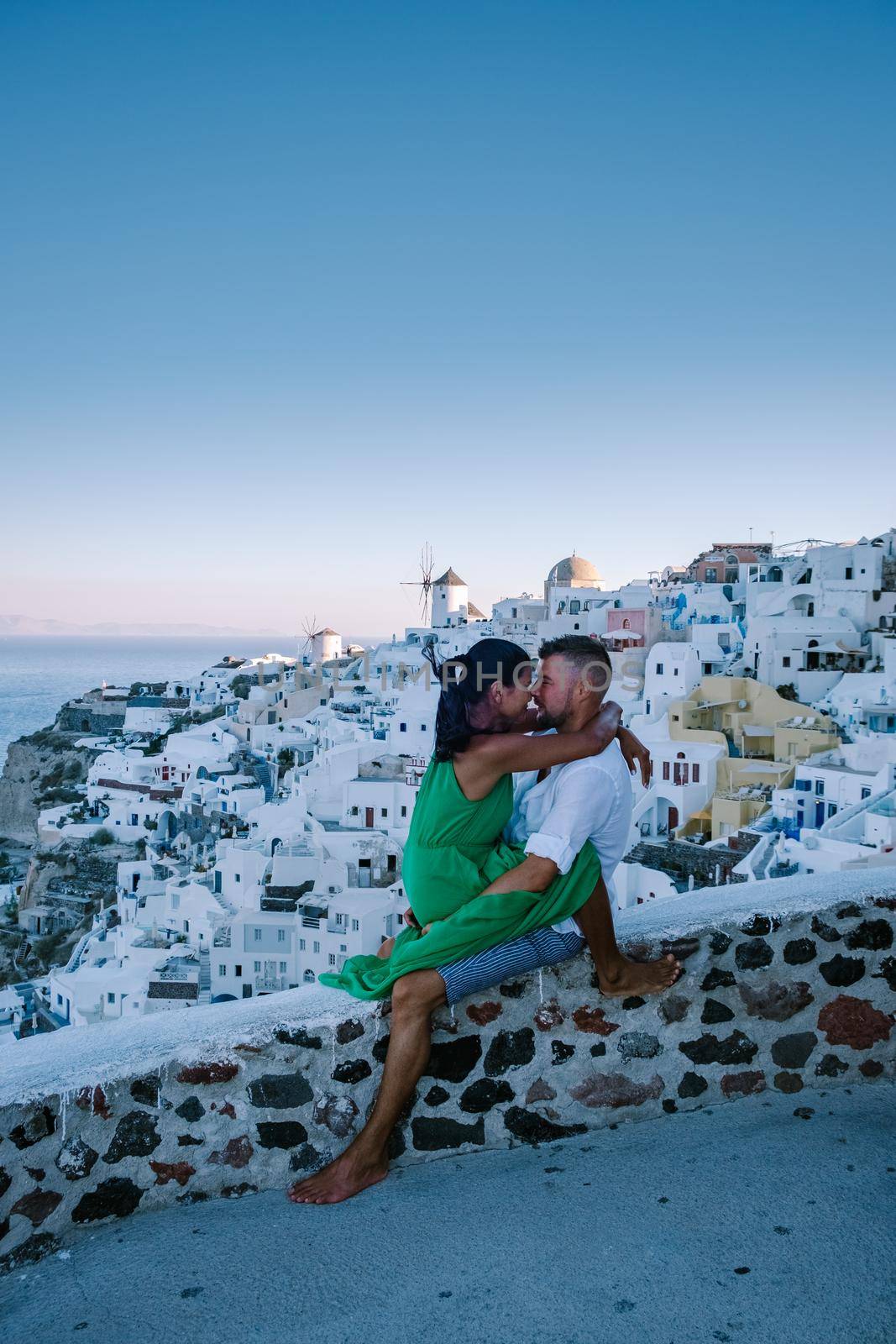 Santorini Greece, young couple on luxury vacation at the Island of Santorini watching sunrise by the blue dome church and whitewashed village of Oia Santorini Greece during sunrise during summer vacation, men and woman on holiday in Greece