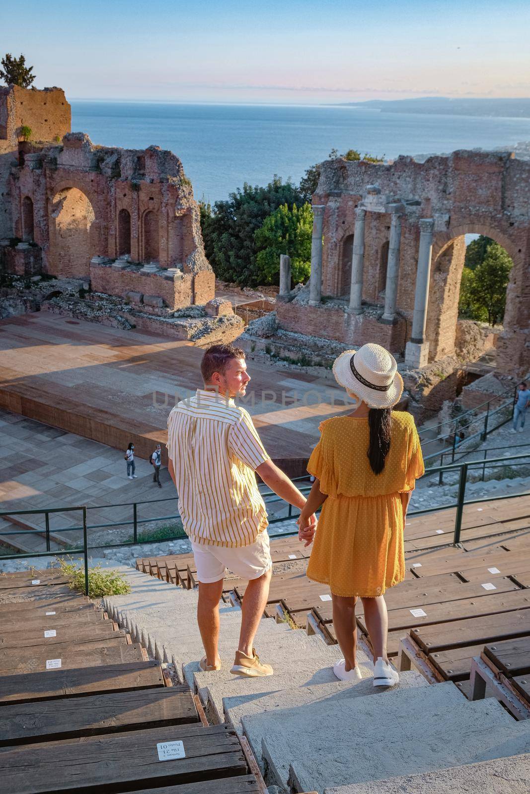 couple men and woman visit Ruins of Ancient Greek theatre in Taormina on background of Etna Volcano, Italy. Taormina located in Metropolitan City of Messina, on east coast of island of Sicily Italy