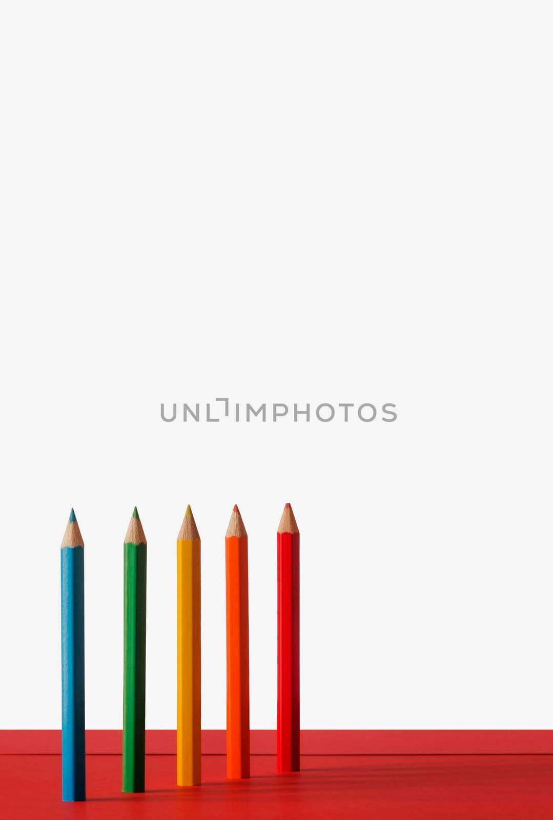 Colored pencils on a white-red background.They are arranged upright sequentially ,they make a slight shadow on the background