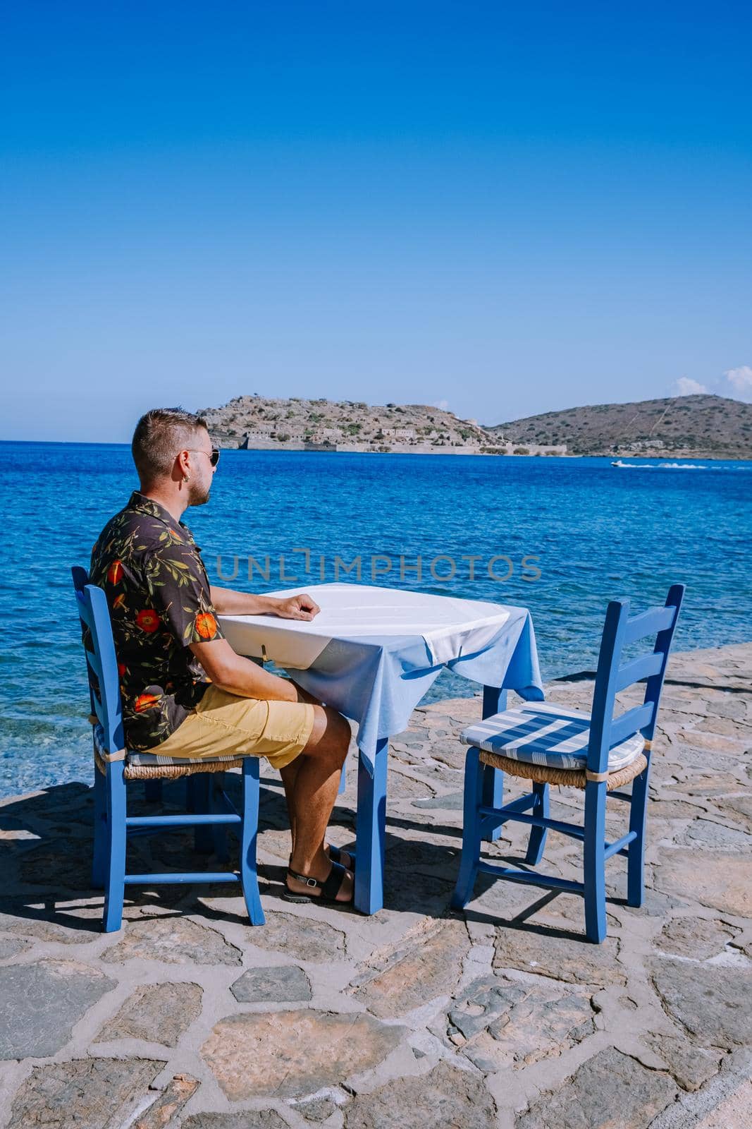 Crete Greece Plaka Lassithi with is traditional blue table and chairs and the beach in Crete Greece. Paralia Plakas, Plaka village Crete, young men mid age having lunch greek taverne