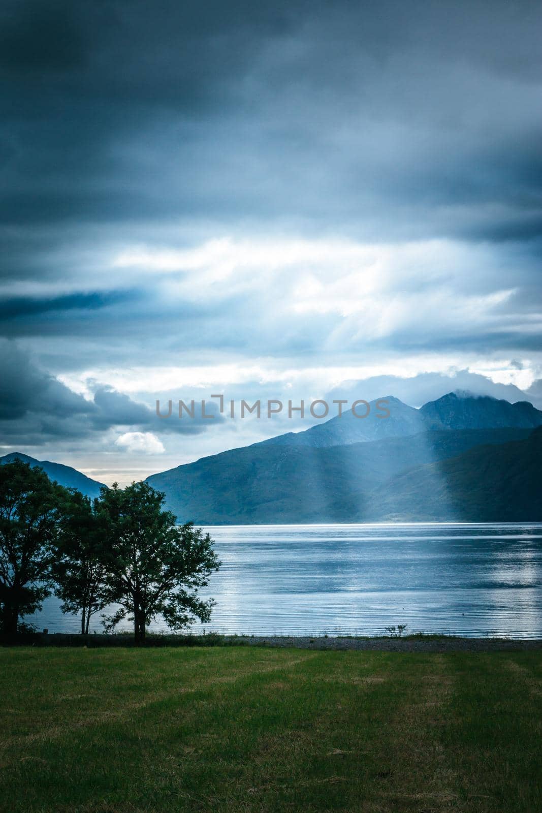 Beautiful mystic landscape lake scenery in Scotland with cloudy sky, meadow, trees and sunbeams