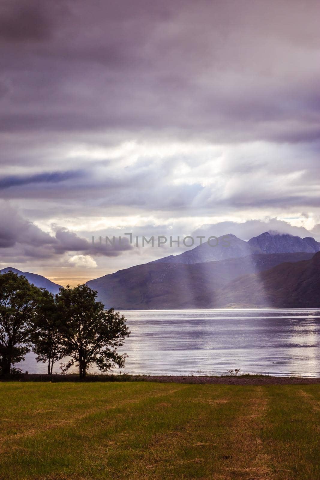 Beautiful mystic landscape lake scenery in Scotland with cloudy sky, meadow, trees and sunbeams
