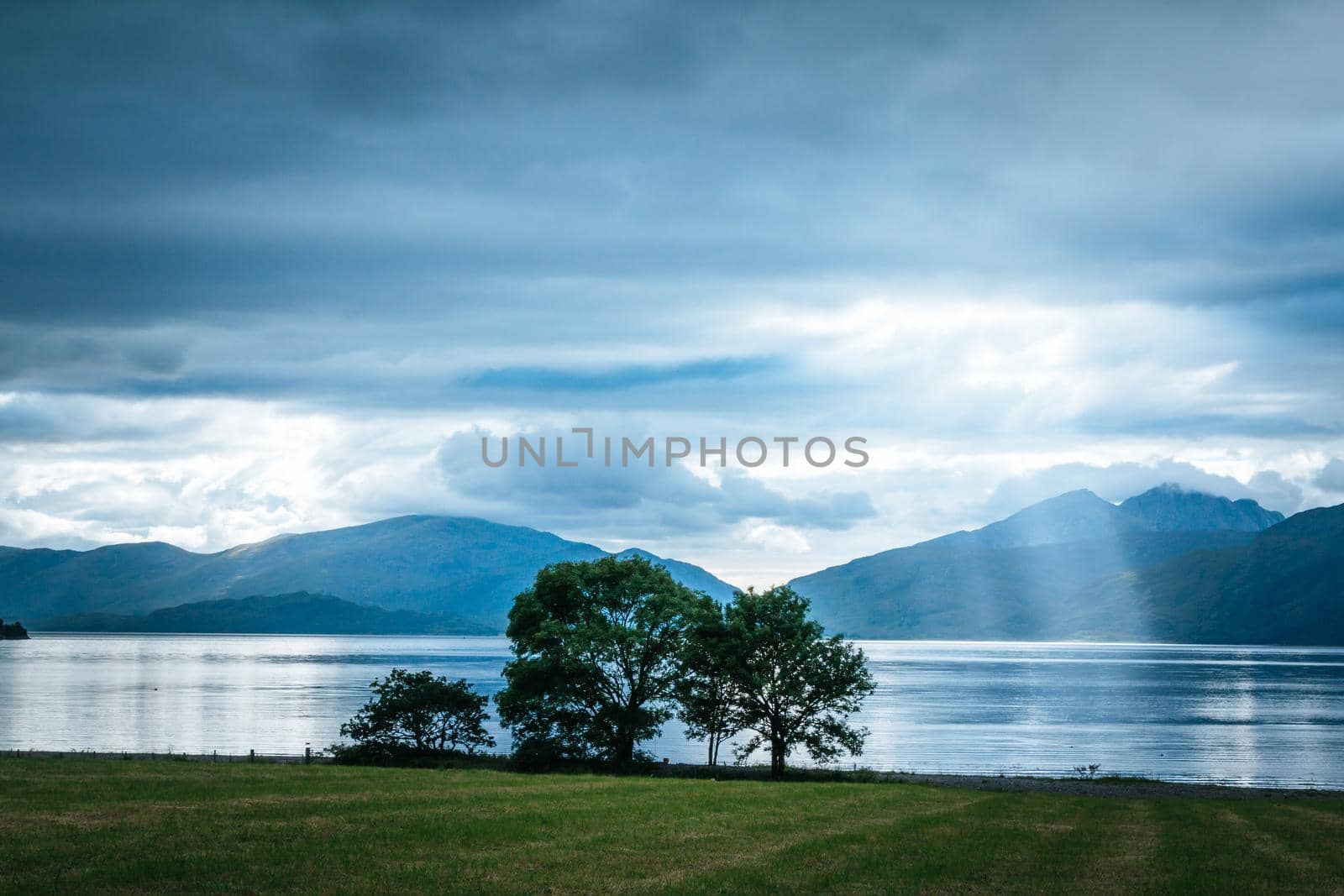 Beautiful mystic landscape lake scenery in Scotland with cloudy sky, meadow, trees and sunbeams