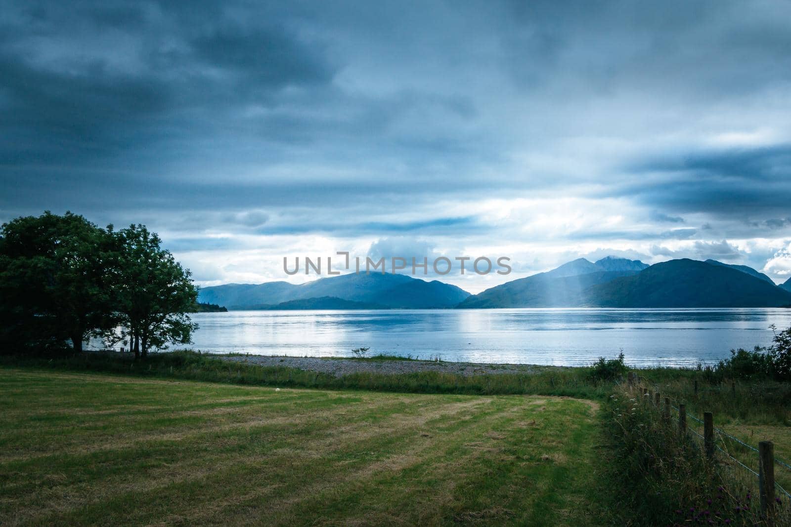 Beautiful mystic landscape lake scenery in Scotland with cloudy sky, meadow, trees and sunbeams