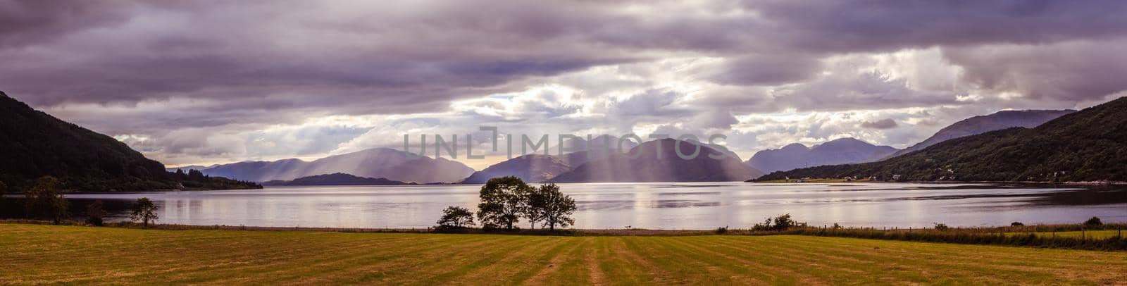 Mystic landscape lake scenery in Scotland: Cloudy sky, meadow, trees and lake with sunbeams, mountain range in the background. Loch Linnhe. by Daxenbichler