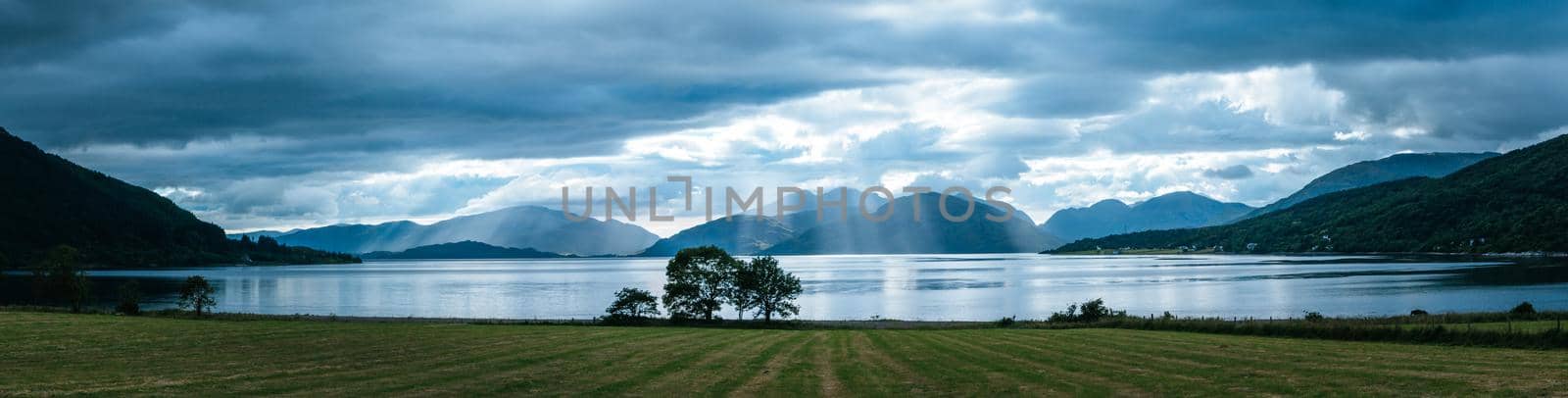 Beautiful mystic landscape lake scenery in Scotland with cloudy sky, meadow, trees and sunbeams