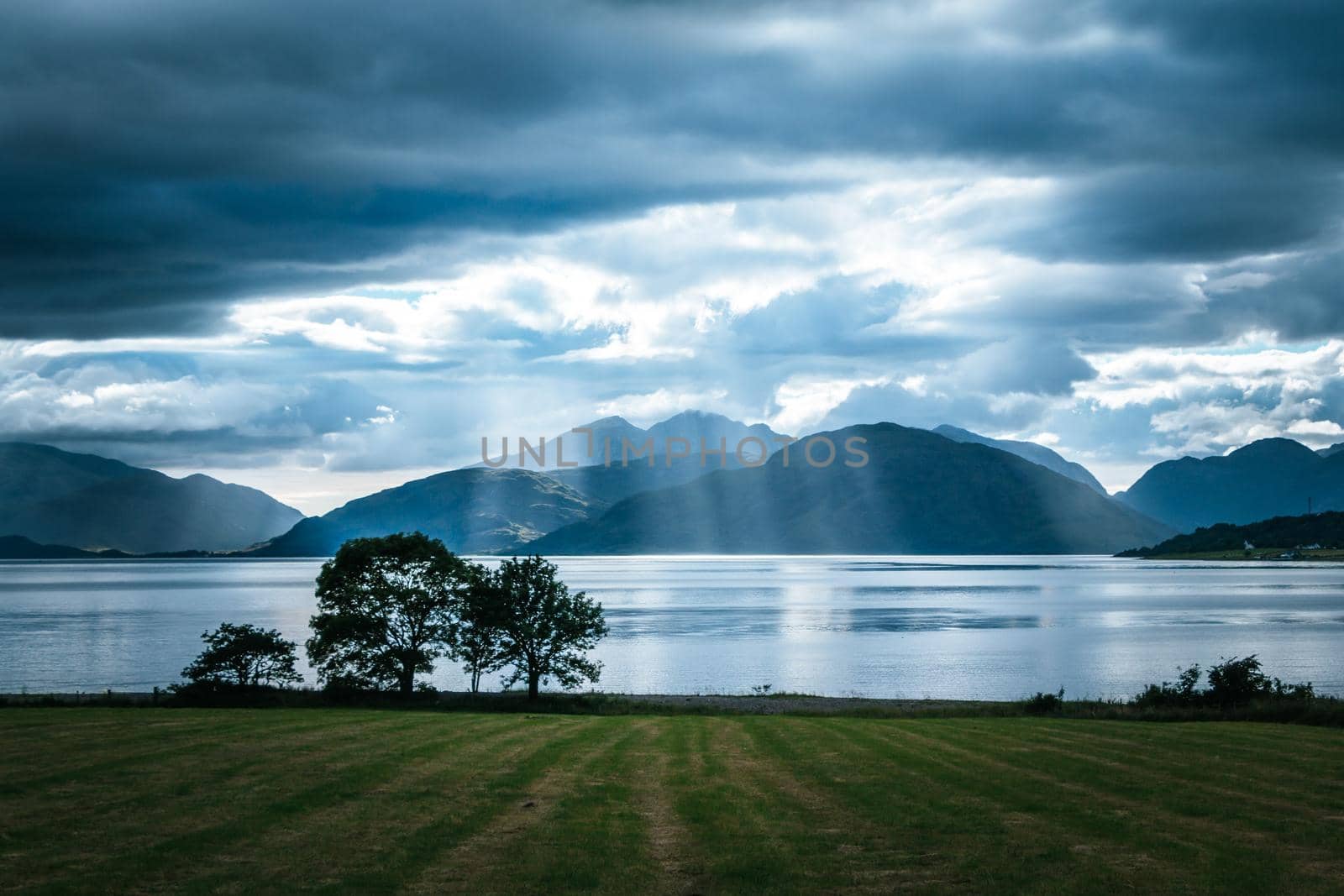 Beautiful mystic landscape lake scenery in Scotland with cloudy sky, meadow, trees and sunbeams