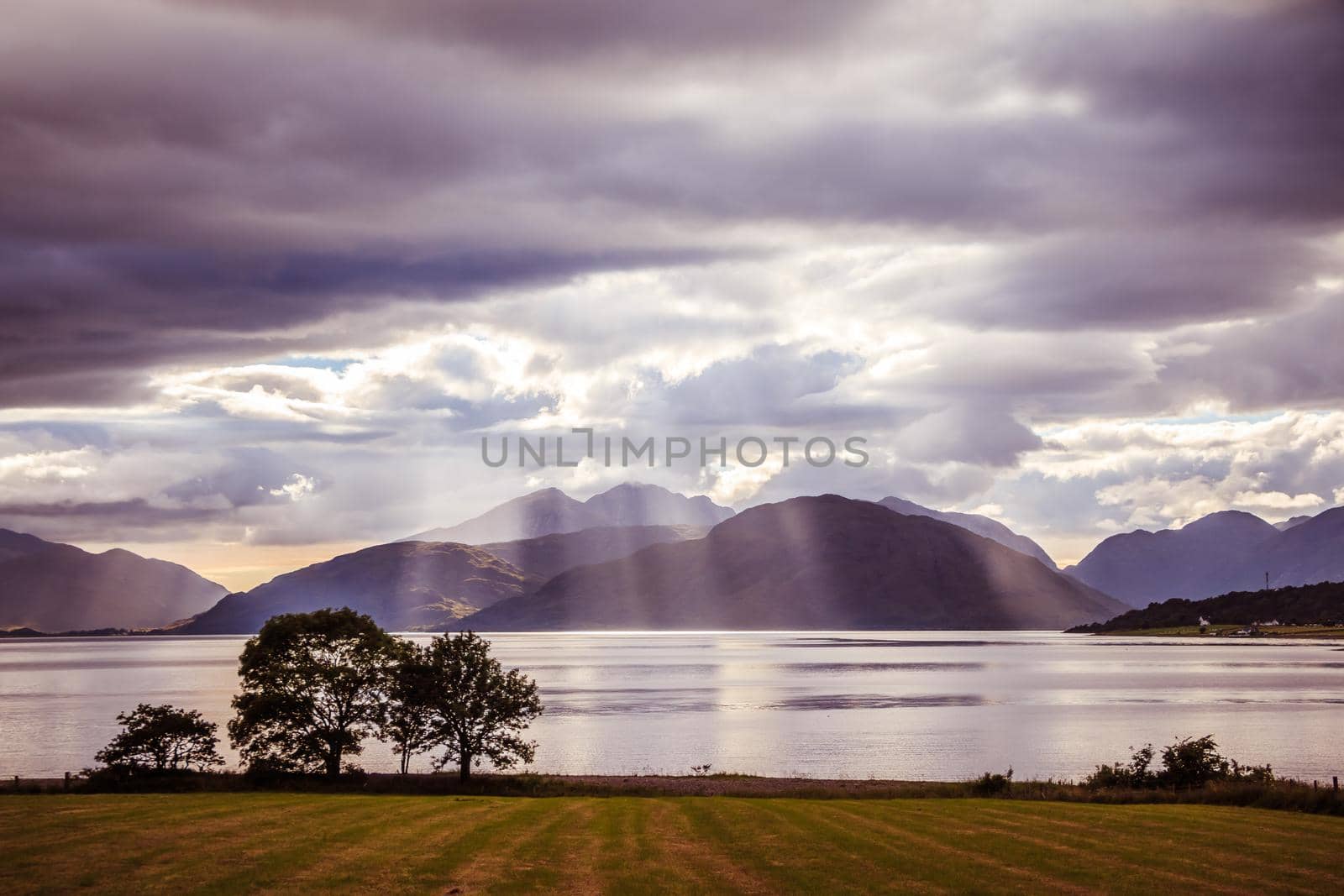 Beautiful mystic landscape lake scenery in Scotland with cloudy sky, meadow, trees and sunbeams