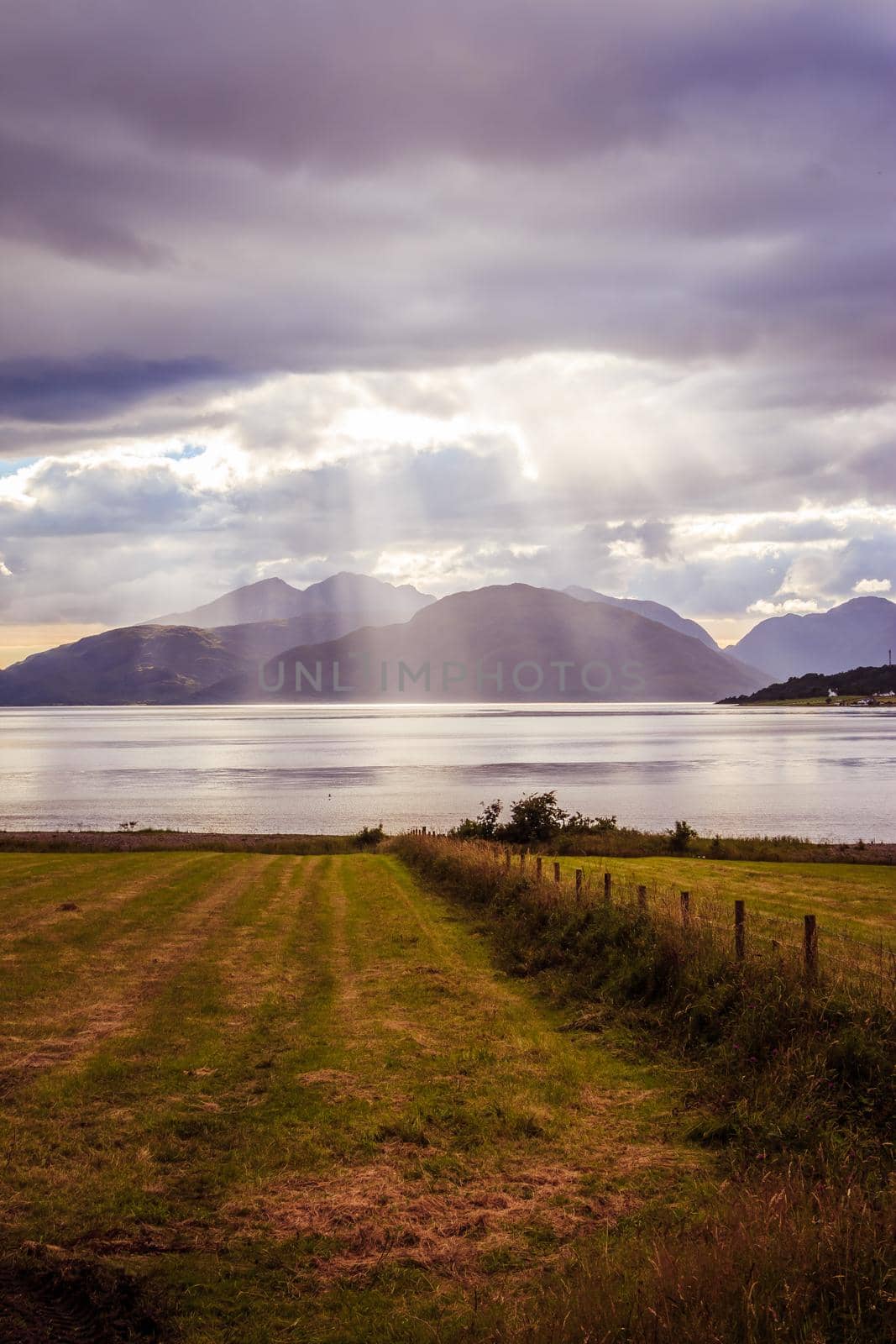 Beautiful mystic landscape lake scenery in Scotland with cloudy sky, meadow, trees and sunbeams