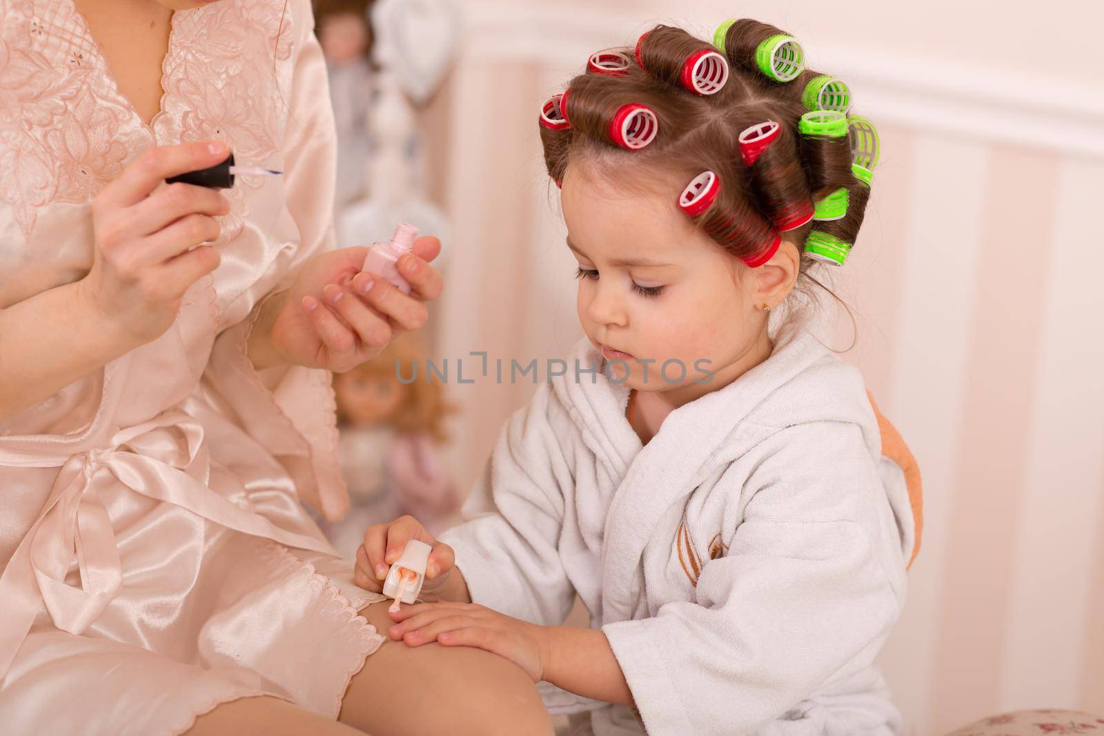 Adorable little girl with her mother in curlers paint their fingernails. Copies mom's behavior. Mom teaches her daughter to take care of herself. Beauty day.