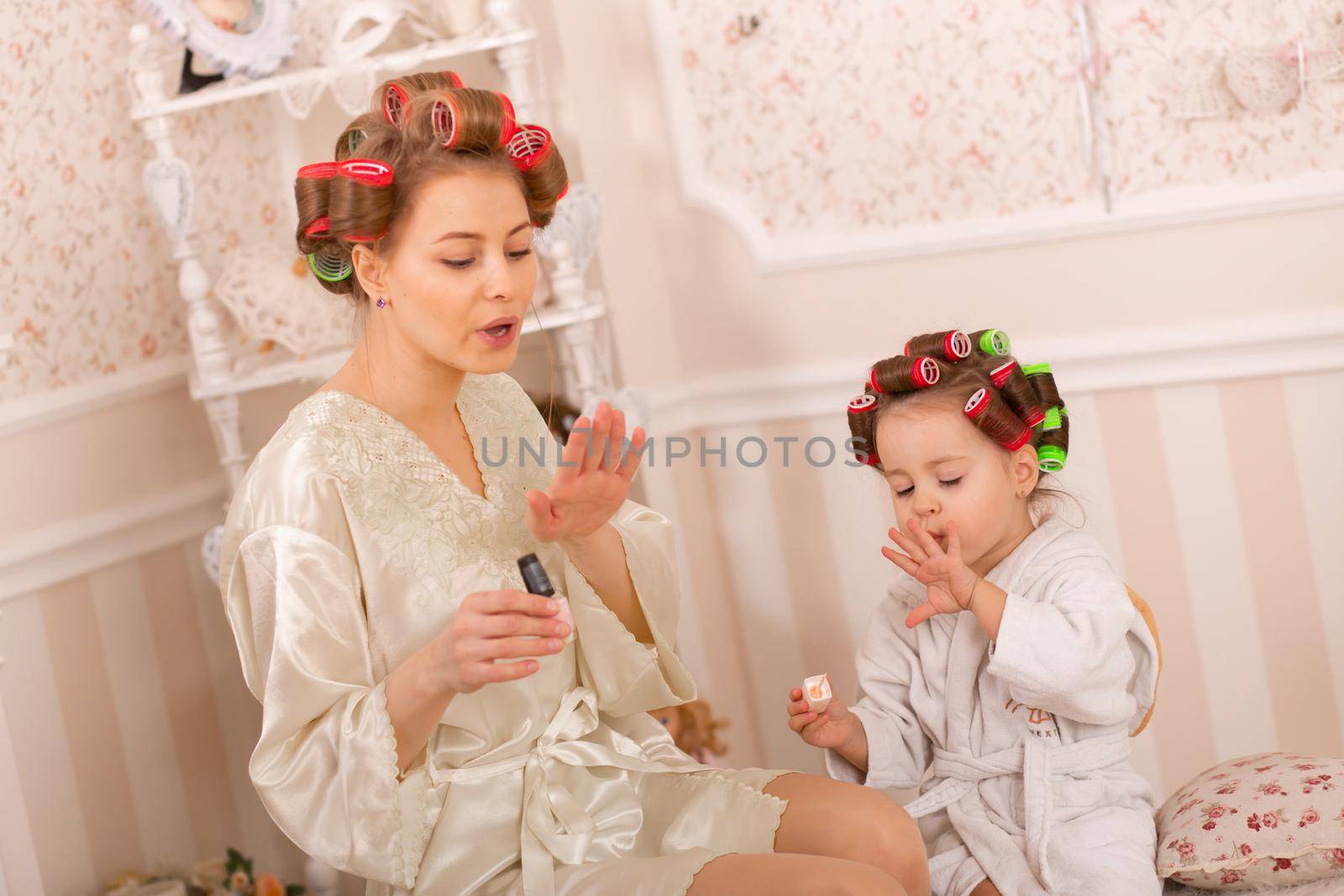Adorable little girl with her mother in curlers paint their fingernails. Copies mom's behavior. Mom teaches her daughter to take care of herself. Beauty day.