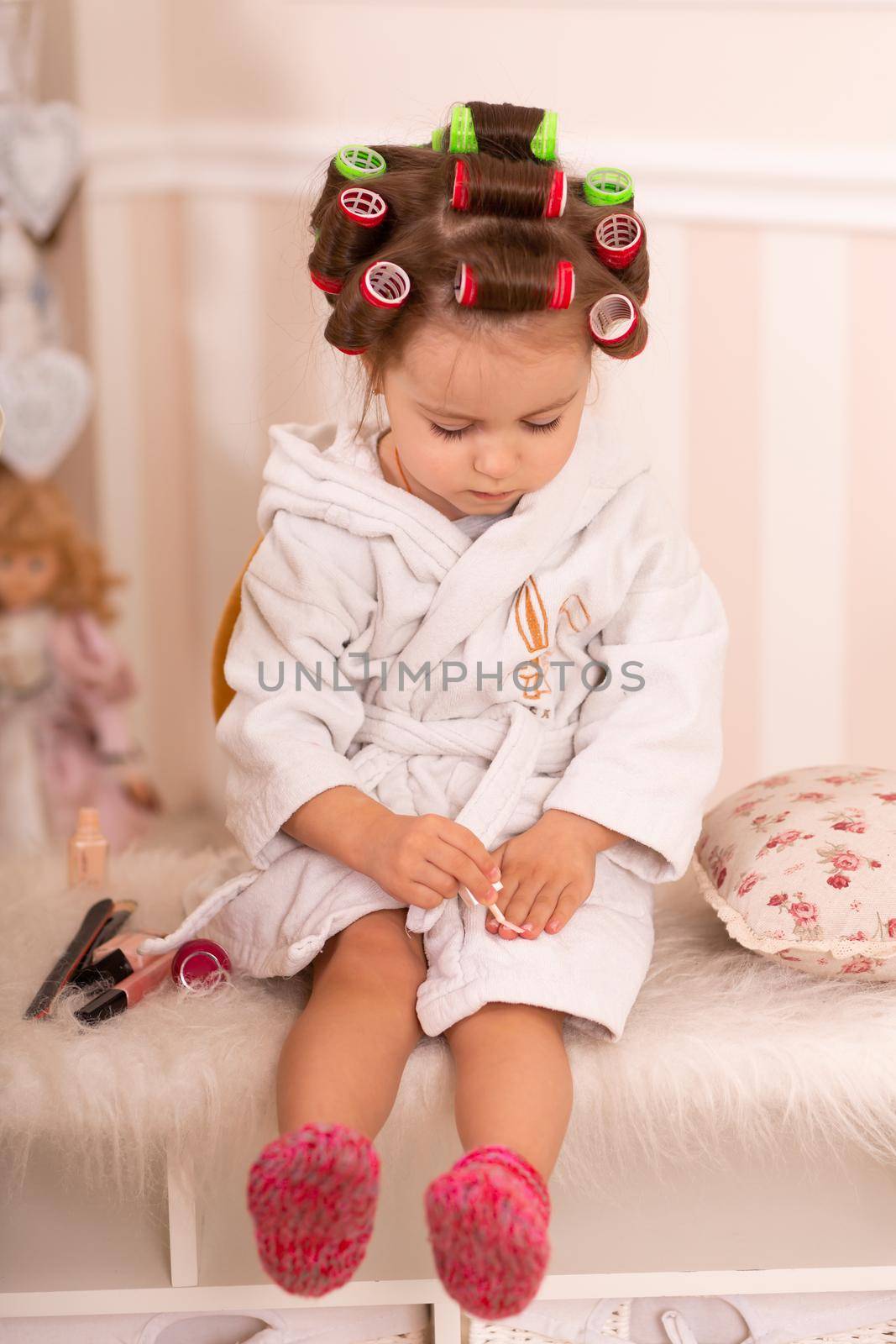 Adorable little girl with curlers paints her fingernails. Copies mom's behavior. Young fashionista. Beauty day.