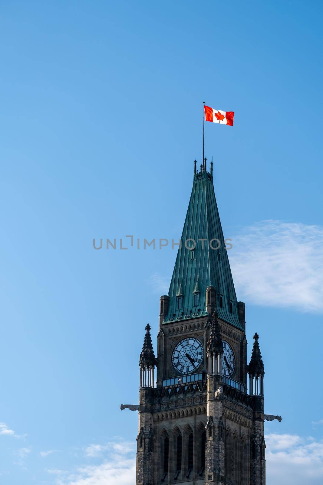 The upper part of the Peace Tower on Parliament Hill in Ottawa, Canada from a low angle against a blue sky with clouds. A Canadian flag flies in the wind on the top of the clock tower.