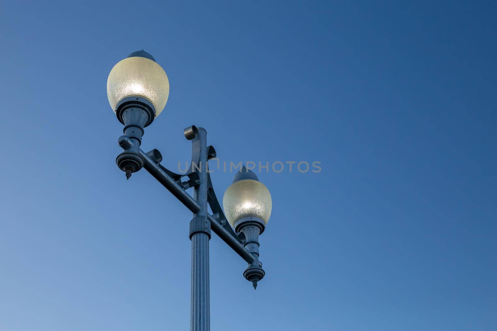 A city lamppost is seen from a low angle against a gradient blue sky. The lights are illuminated in foggy glass casing atop a blue metal stand.