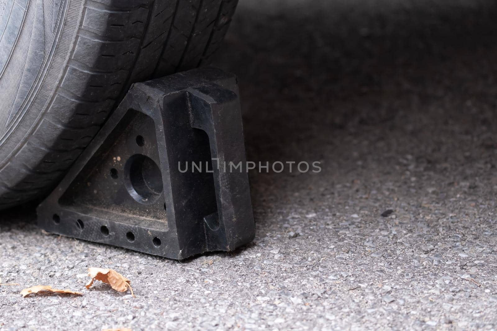 A black rubber wheel chock on the ground is placed behind a car tire to keep the vehicle in place. It is on a paved asphalt driveway with autumn leaves beside it.